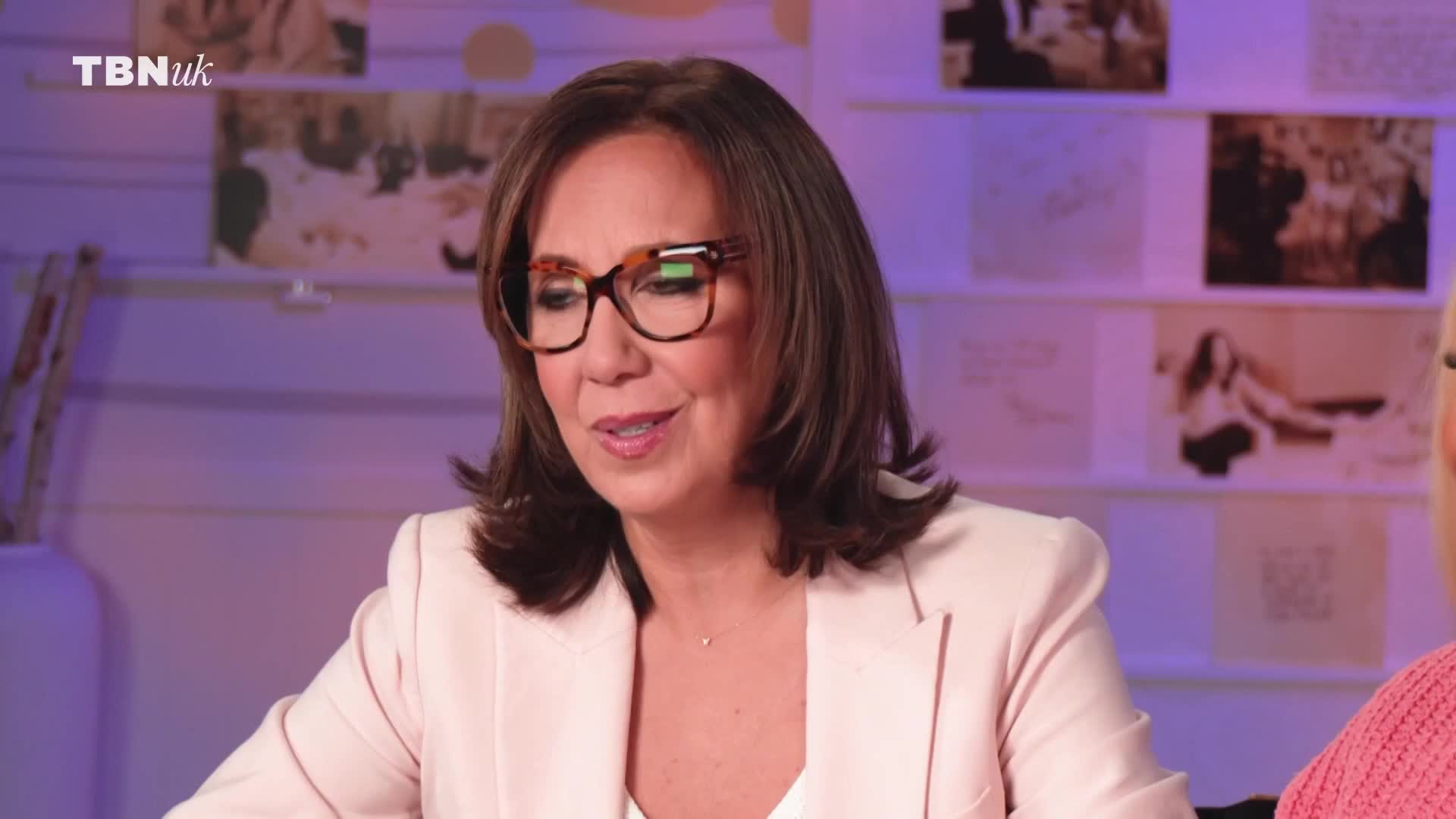 A woman with tortoiseshell glasses leans forward, speaking. Behind her, a TBN UK logo sits in the corner of a wall adorned with framed pictures and sketches.