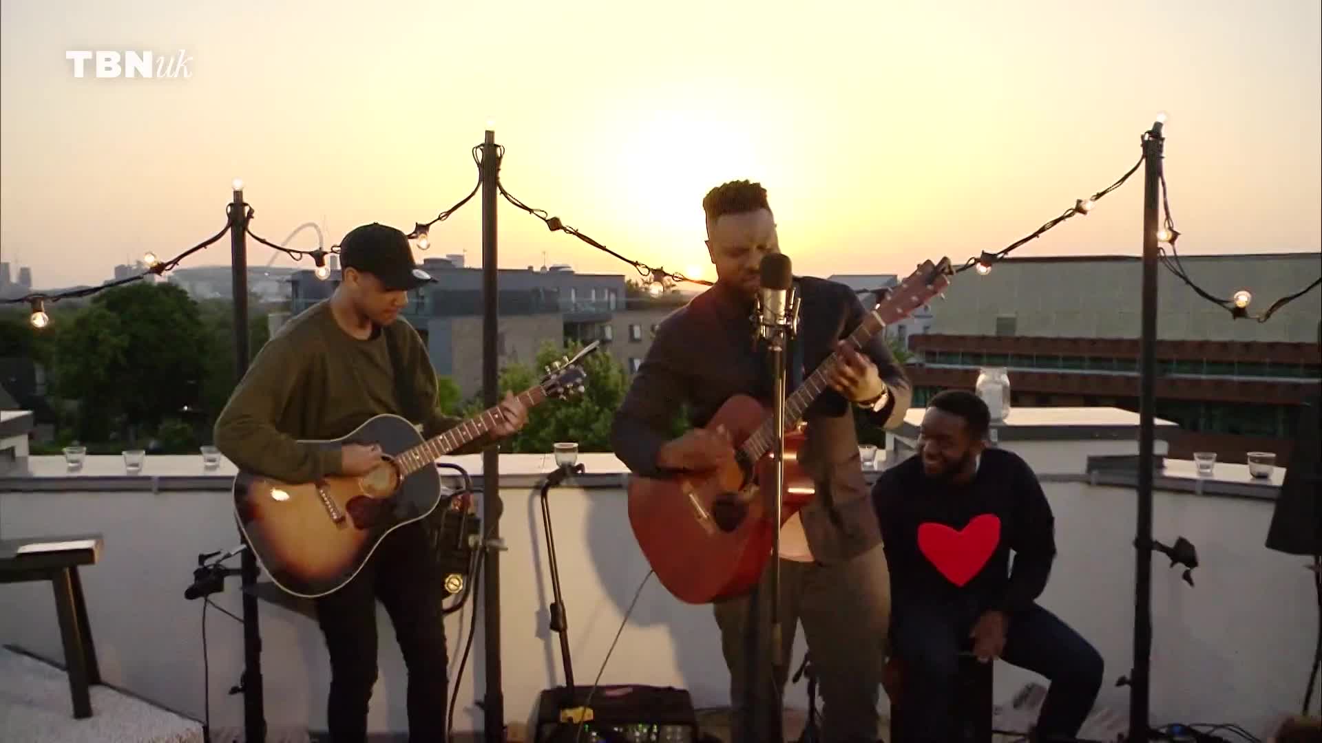 Three men are playing guitars on a rooftop as the sun sets over the city. The TBN UK logo is visible in the corner of the screen. Three men are playing guitars on a rooftop as the sun sets over the city. The TBN UK logo is visible in the corner of the screen.