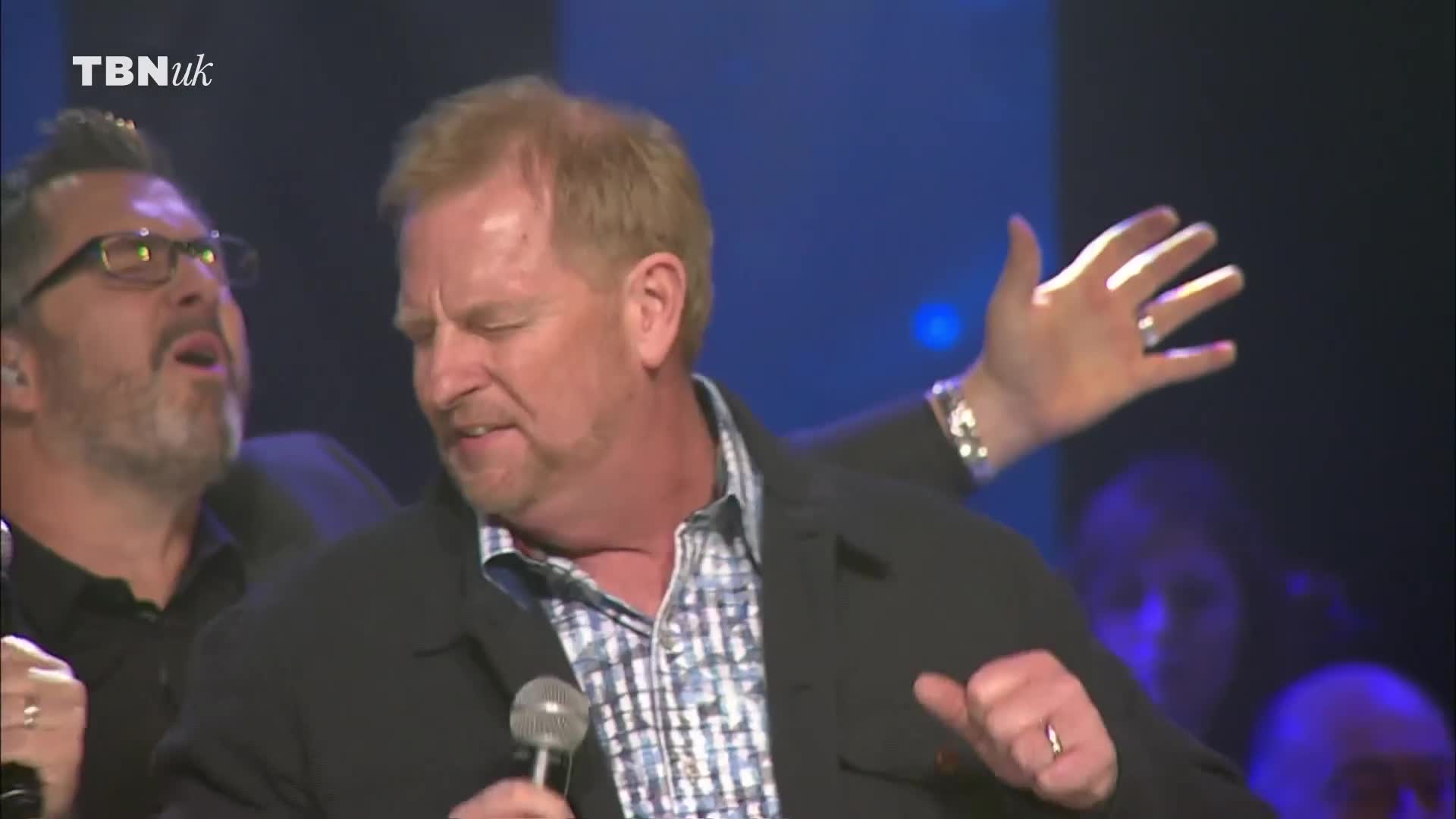 A man with a microphone sings with his eyes closed, his hand raised in the air. Another man beside him, wearing glasses, sings with his mouth open. A man with a microphone sings with his eyes closed, his hand raised in the air. Another man beside him, wearing glasses, sings with his mouth open.