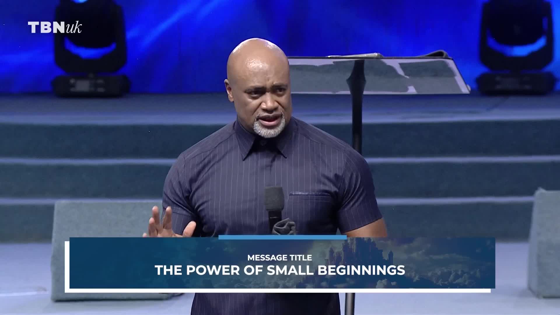 A man with a shaved head and a goatee speaks into a microphone, his hands gesturing as he addresses an unseen audience. A banner below him reads "THE POWER OF SMALL BEGINNINGS," with the TBN UK logo visible in the corner. A man with a shaved head and a goatee speaks into a microphone, his hands gesturing as he addresses an unseen audience. A banner below him reads "THE POWER OF SMALL BEGINNINGS," with the TBN UK logo visible in the corner.
