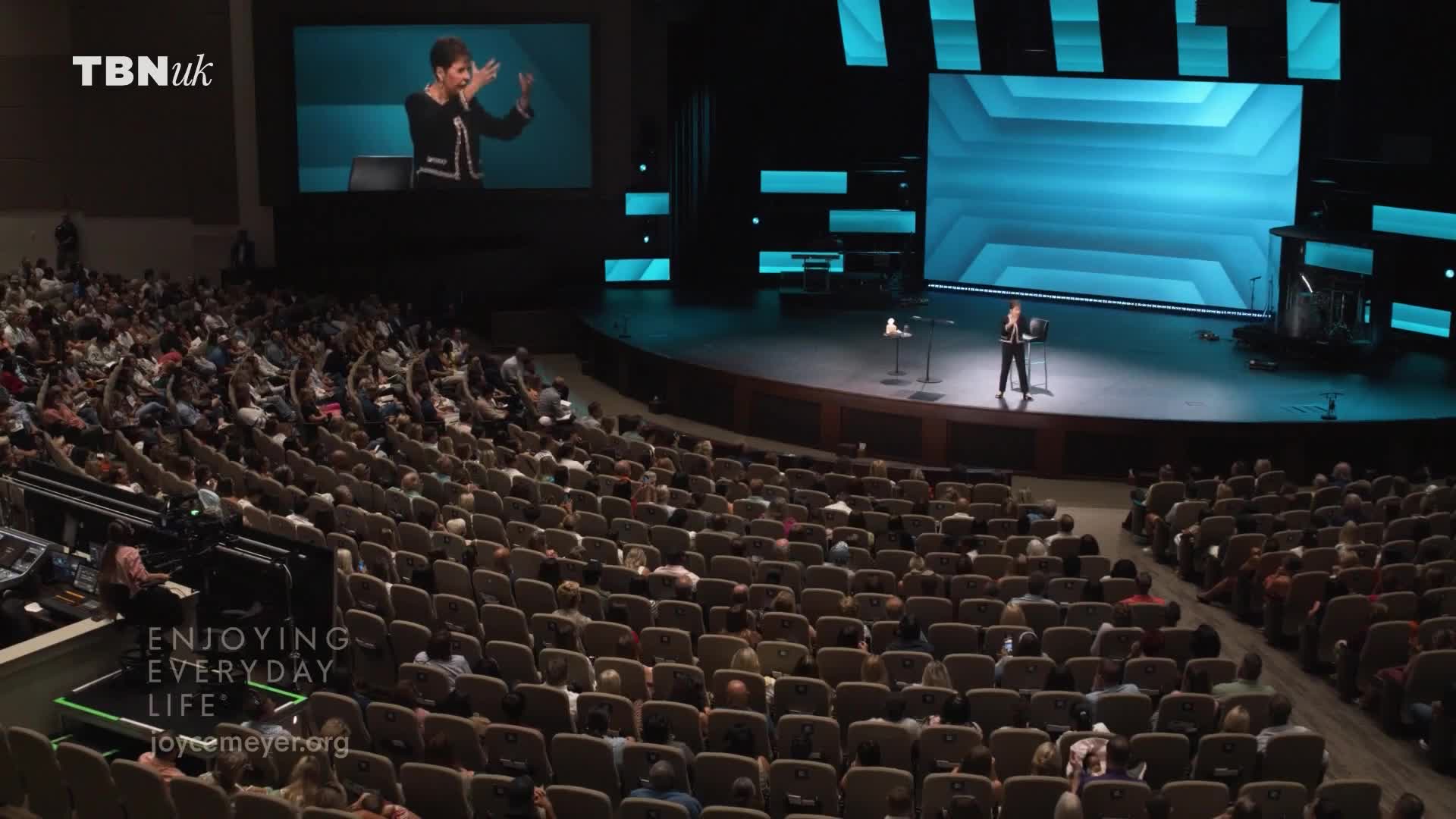 A woman stands on a stage, speaking to a large audience seated in a theatre. A screen behind her shows a live feed of her speaking, with the TBN UK logo in the corner. A woman stands on a stage, speaking to a large audience seated in a theatre. A screen behind her shows a live feed of her speaking, with the TBN UK logo in the corner.