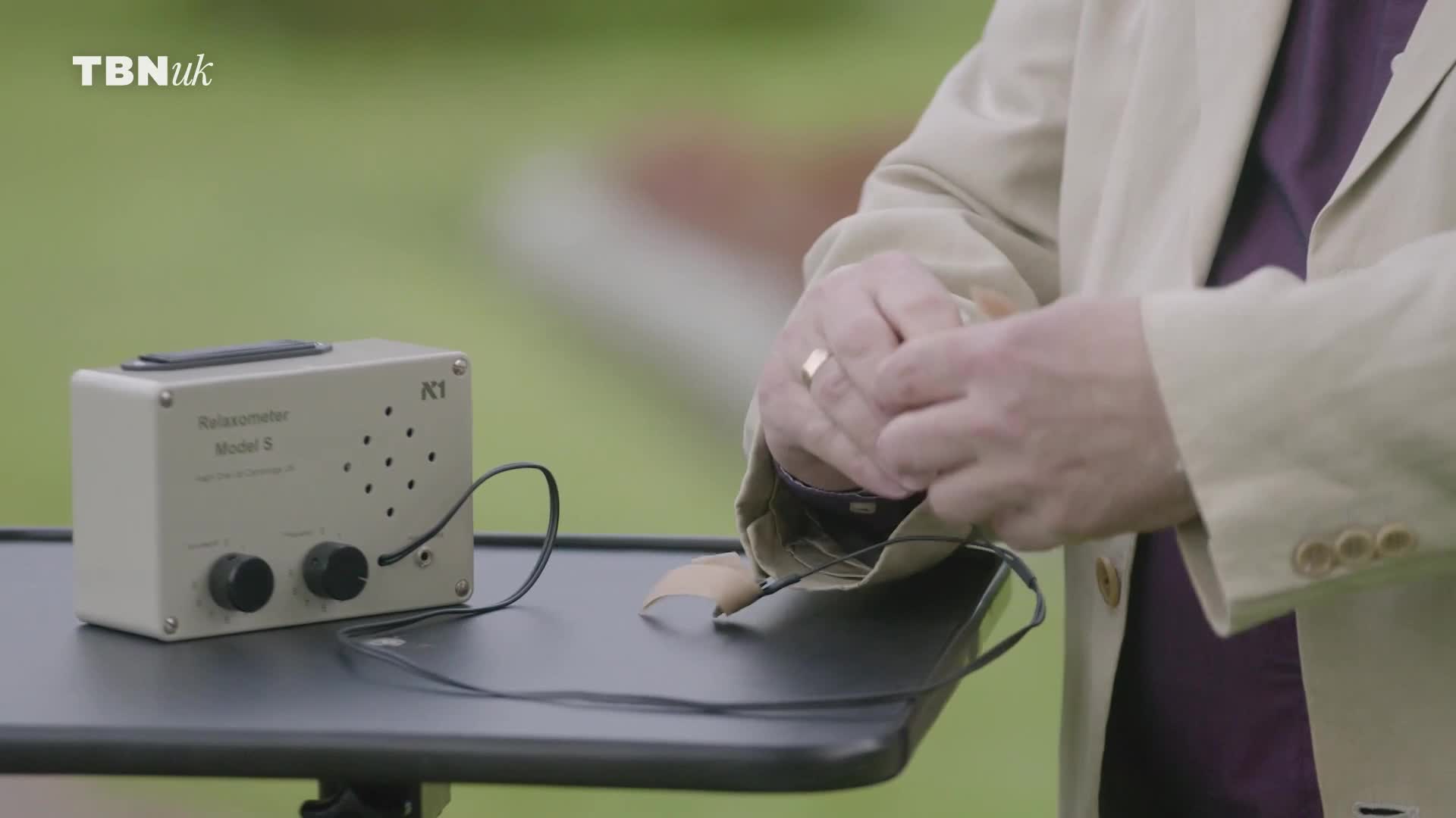 A man in a beige jacket is carefully adjusting a small, beige device with two knobs and a speaker grille. A thin wire snakes from the device, disappearing under his jacket. A man in a beige jacket is carefully adjusting a small, beige device with two knobs and a speaker grille. A thin wire snakes from the device, disappearing under his jacket.