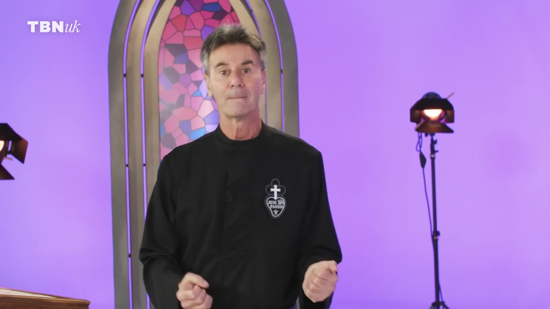 A man in a black shirt with a crest stands before a stained-glass-like backdrop. A TBN UK logo appears in the upper left corner. A man in a black shirt with a crest stands before a stained-glass-like backdrop. A TBN UK logo appears in the upper left corner.