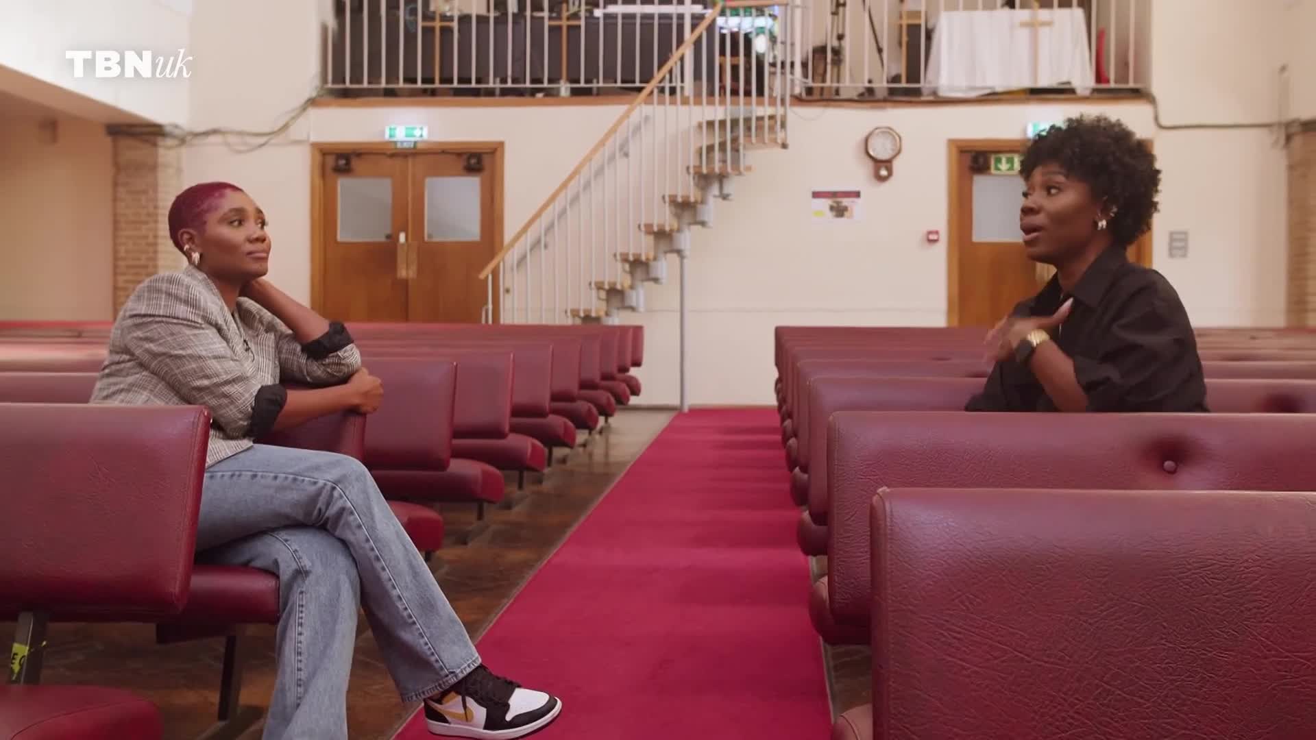 Two women sit in a church, facing each other across a red carpeted aisle. The woman on the left, with short pink hair, leans back with her hand behind her head. The woman on the right, with dark curly hair, gestures with her hands as she speaks. Two women sit in a church, facing each other across a red carpeted aisle. The woman on the left, with short pink hair, leans back with her hand behind her head. The woman on the right, with dark curly hair, gestures with her hands as she speaks.