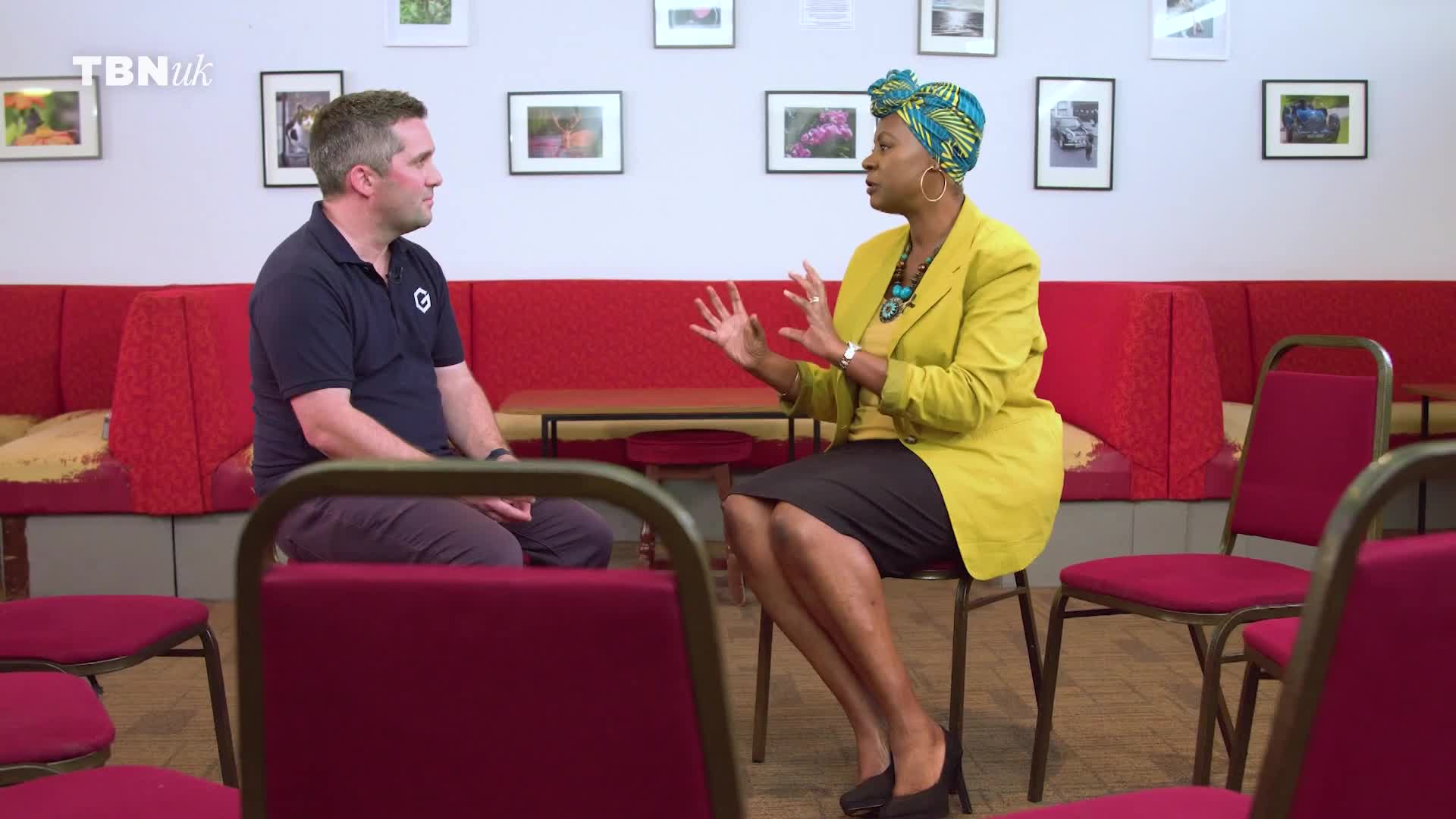 A man in a dark polo shirt sits facing a woman in a bright yellow jacket and headscarf. They are in a room with red seating and framed pictures on the wall. A man in a dark polo shirt sits facing a woman in a bright yellow jacket and headscarf. They are in a room with red seating and framed pictures on the wall.