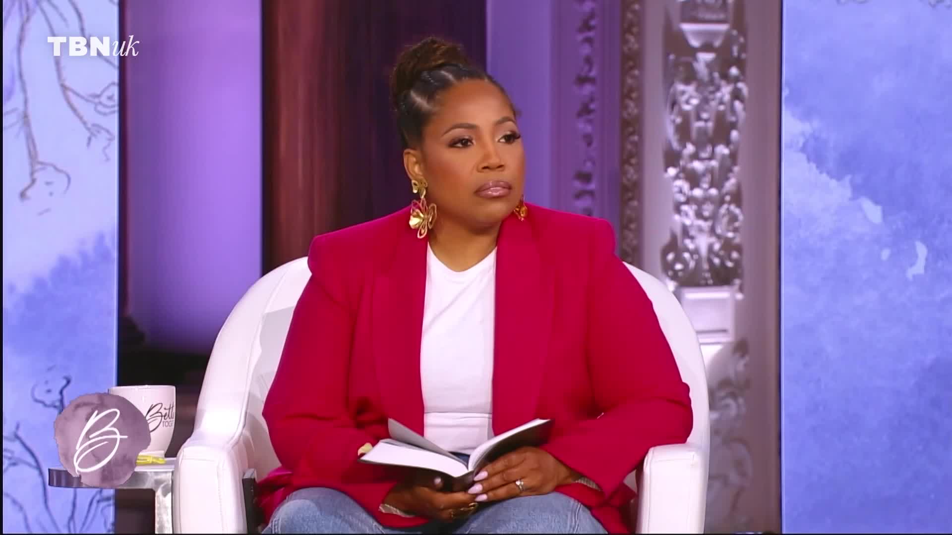 A woman in a bright pink blazer sits in a white chair, holding a small book. The TBN UK logo appears in the top left corner of the screen. A woman in a bright pink blazer sits in a white chair, holding a small book. The TBN UK logo appears in the top left corner of the screen.