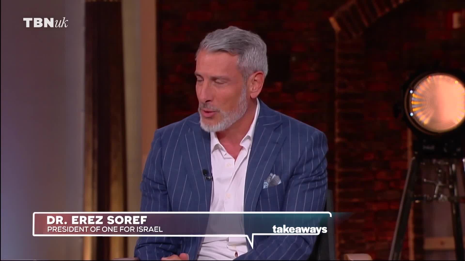 A man with grey hair and a beard, wearing a pinstripe suit, speaks into a microphone. The TBN UK logo appears in the top left corner of the screen.