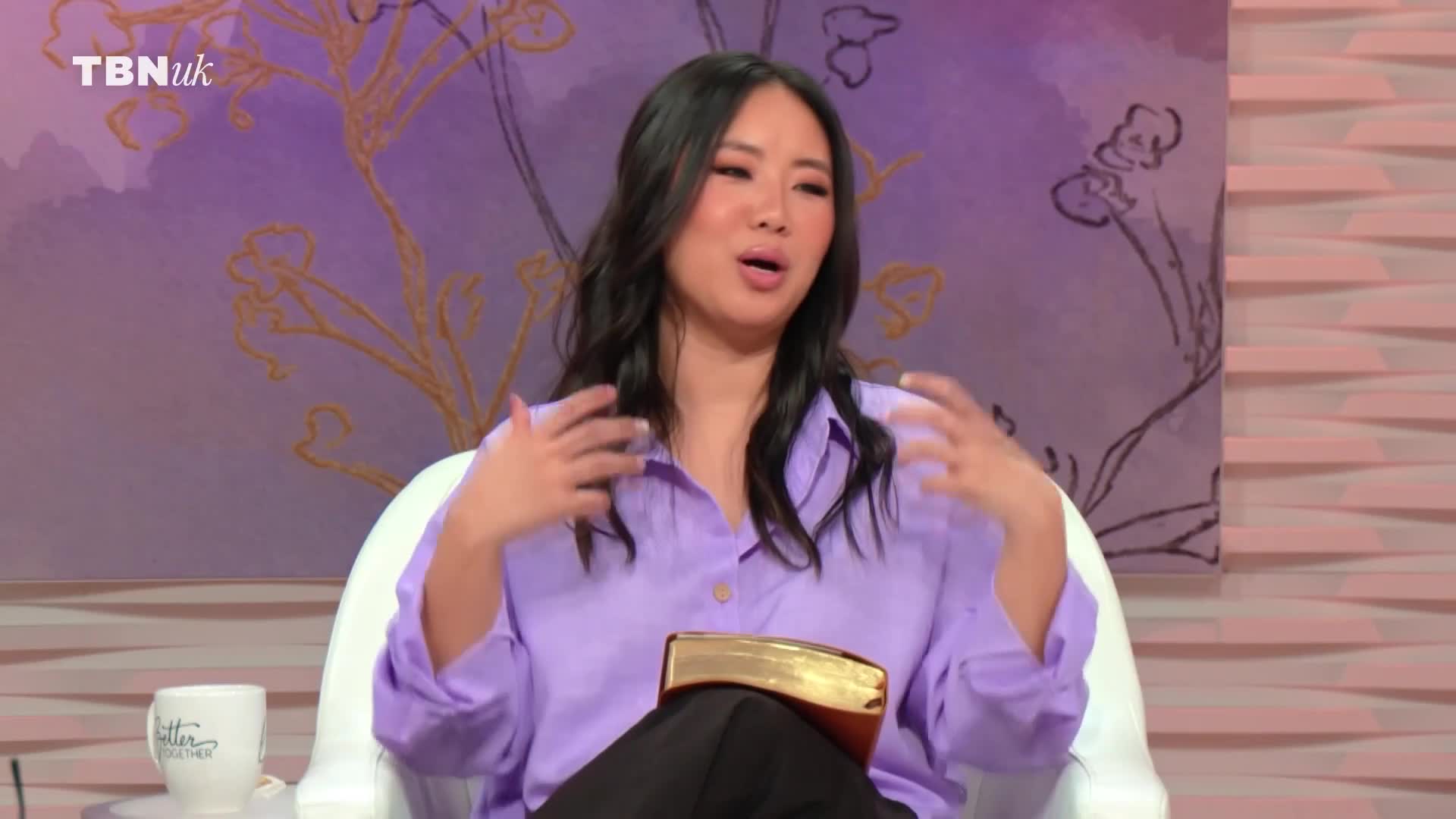 A woman in a lilac shirt gestures with both hands while speaking. A golden-bound book rests open on her lap.