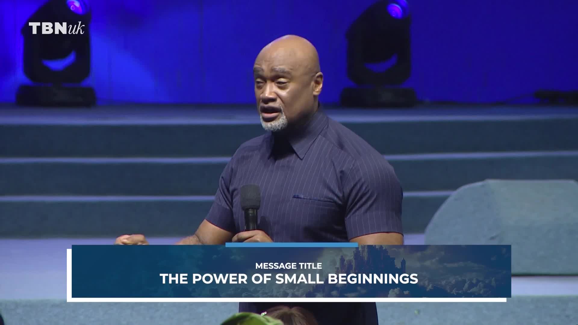 A man in a dark blue, pinstriped shirt holds a microphone and speaks with his left hand extended. The TBN UK logo appears in the upper left corner of the screen.