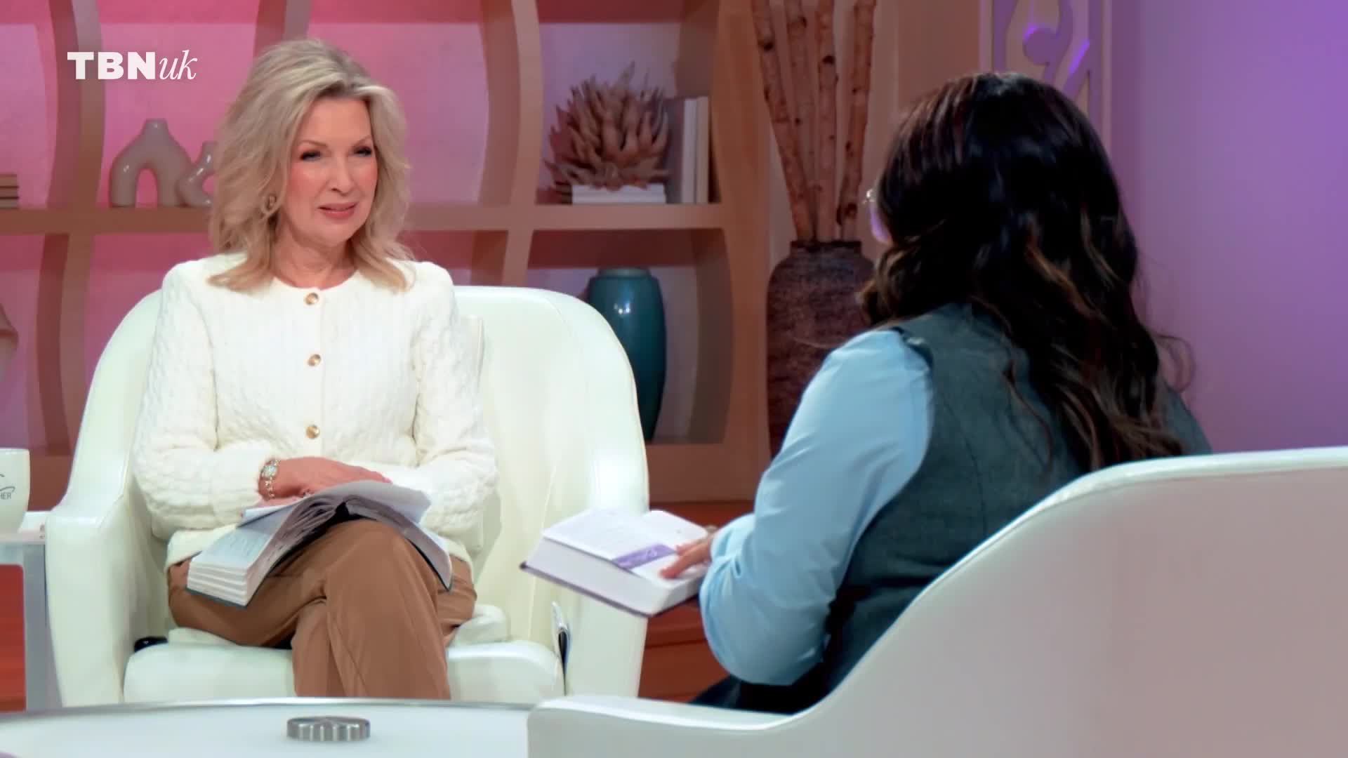 Two women sit across from each other on a television set, each with a book open in their lap. The woman on the left, wearing a cream cable-knit cardigan, smiles as she speaks. The woman on the right, in a blue shirt and grey vest, turns her head slightly as she listens.