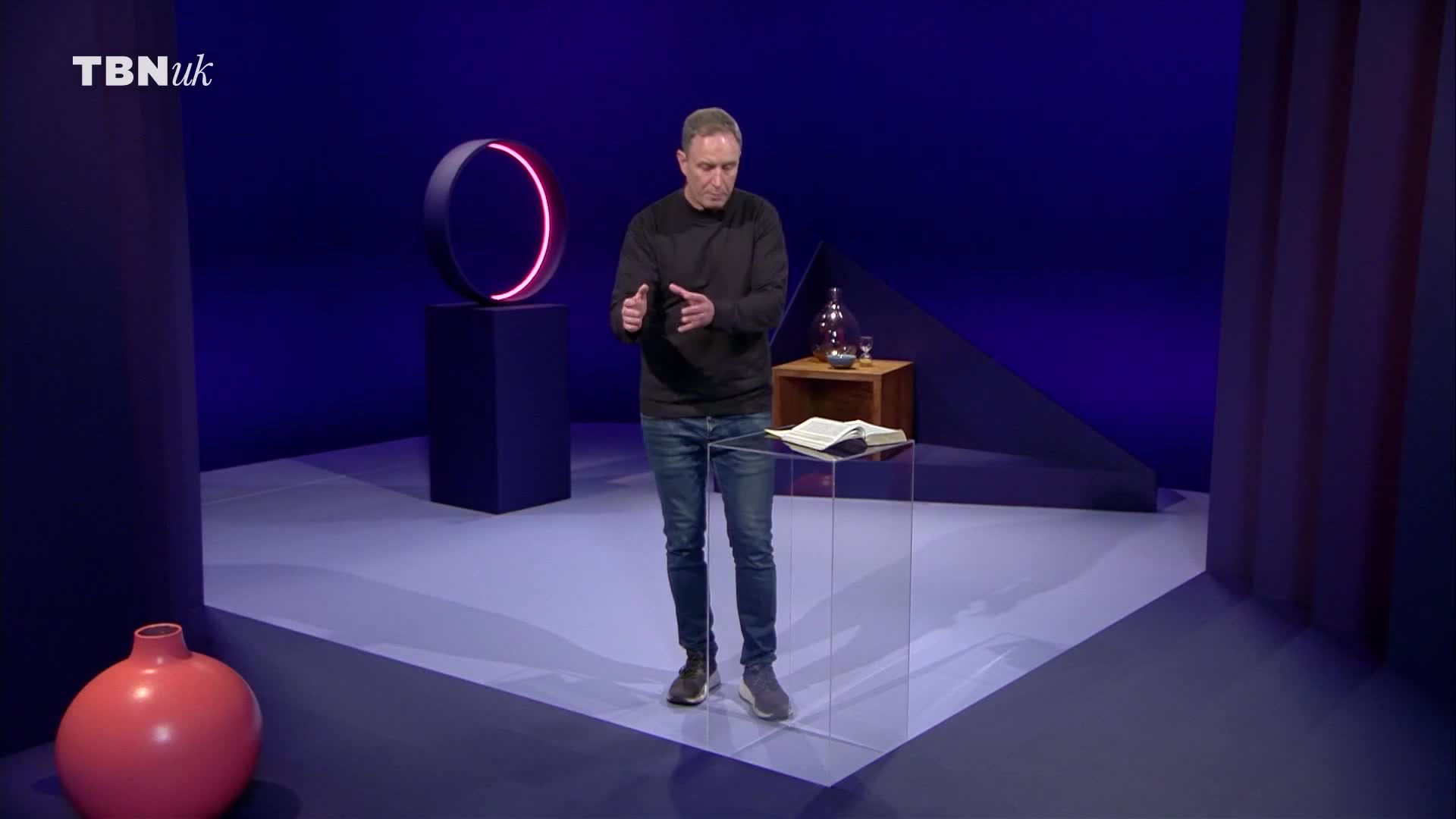 A man in a black sweater and jeans stands on a stage, gesturing with his hands. On a clear podium before him rests an open book, and to his right, a small wooden table holds a glass decanter and a goblet.
