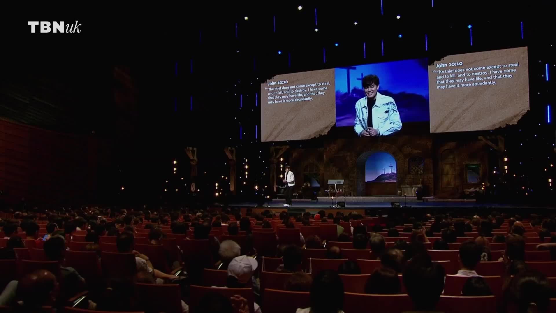 A young man in a white jacket stands on a stage, speaking to a large audience. Behind him, a massive screen displays scripture and a close-up of his face. The TBN UK logo is visible in the top left corner.
