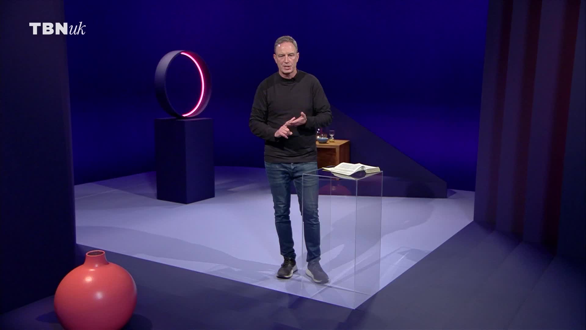 A man stands in a studio, gesturing with his hands as he speaks. A TBN UK logo is visible in the top left corner.