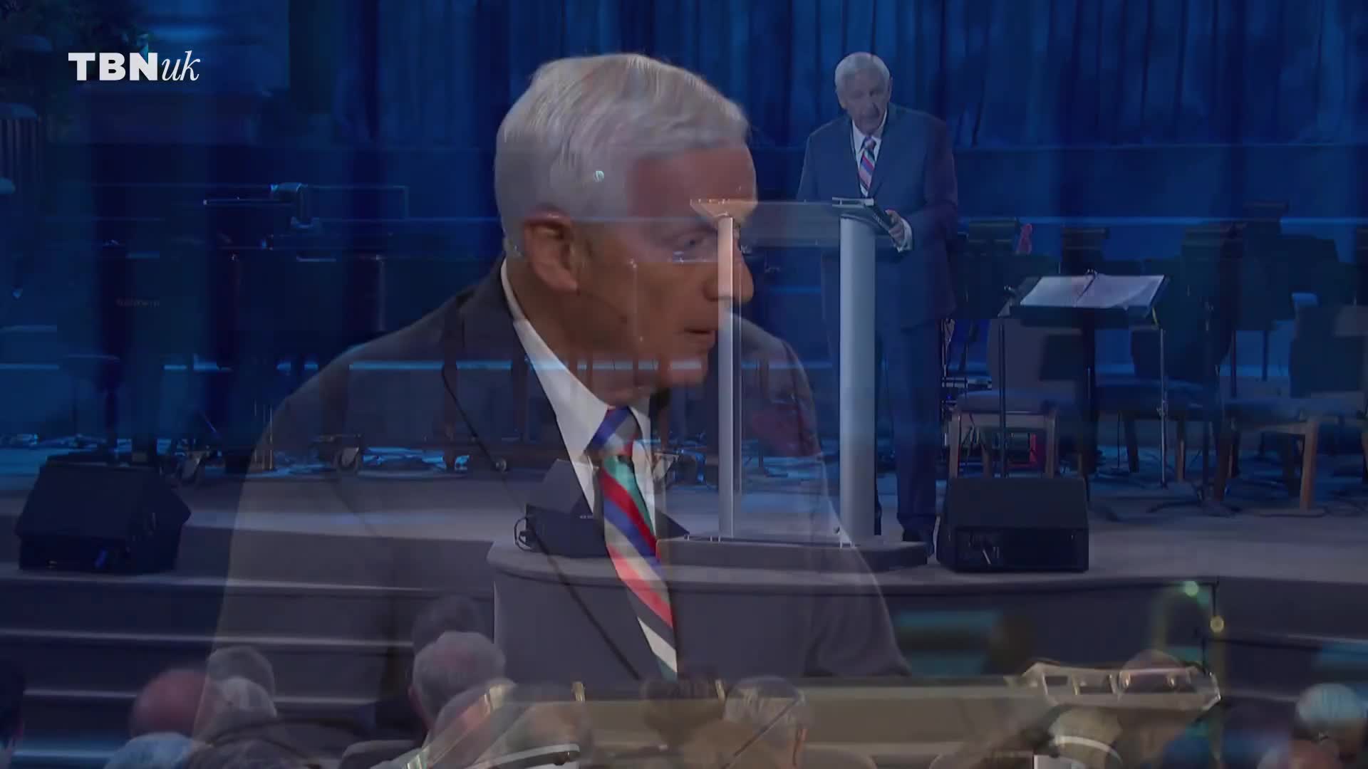 A man in a suit stands at a podium, his gaze fixed forward. Behind him, another man in a suit reads from a paper at a different lectern.