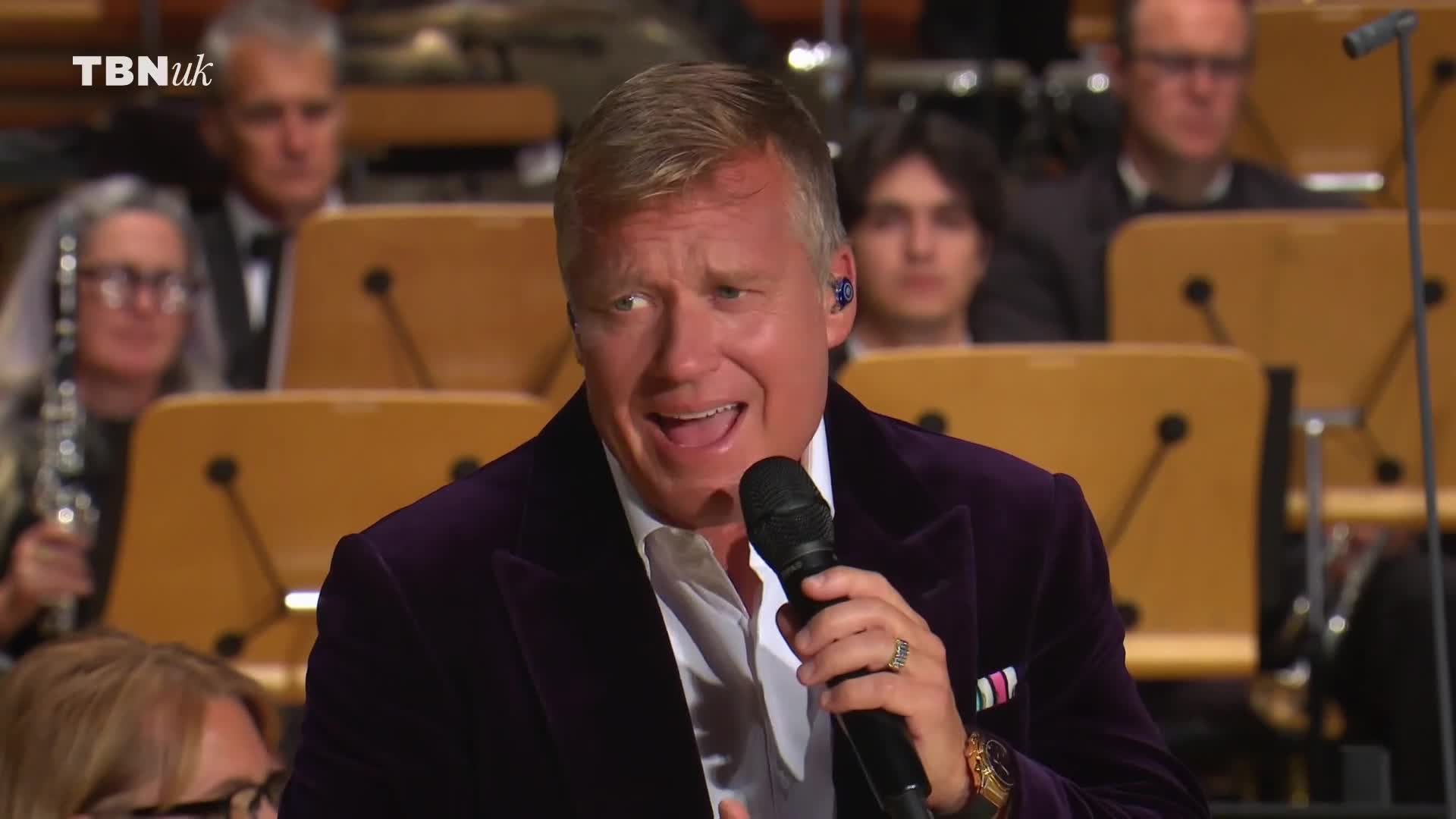 A man in a purple velvet jacket sings into a microphone, his mouth open mid-word. Behind him, an orchestra is seated, their instruments ready.