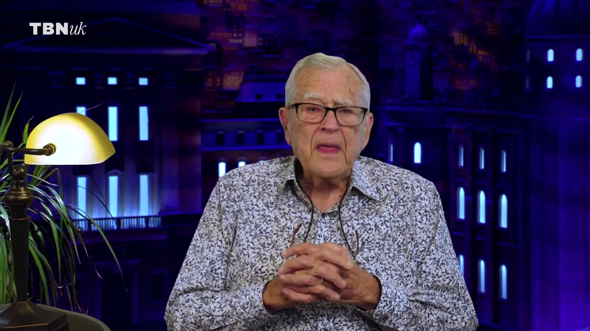 An older gentleman with glasses is speaking, his hands clasped in front of him. Behind him, a backdrop depicts a stylized building with illuminated windows, the TBN UK logo visible in the corner.