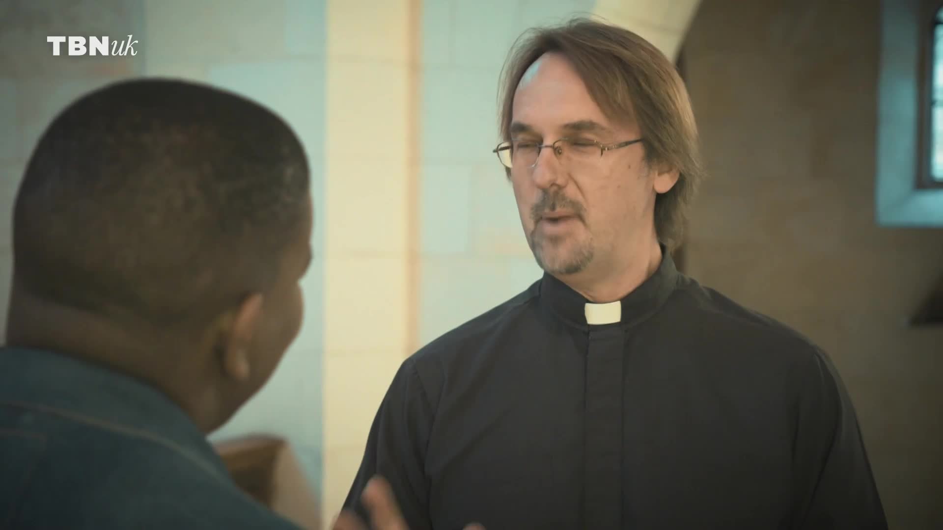 A man in a black clerical shirt with a white collar speaks to another man, his hands gesturing slightly. The TBN UK logo is visible in the corner, hinting at a broadcast from the United Kingdom.
