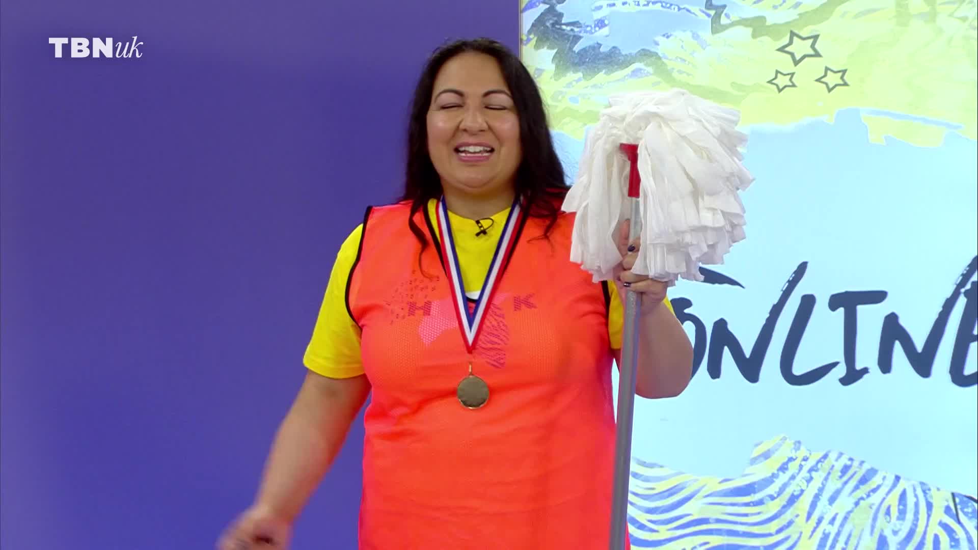 A woman in a bright orange vest and yellow shirt stands holding a mop. She wears a medal around her neck, and the TBN UK logo is visible in the corner.