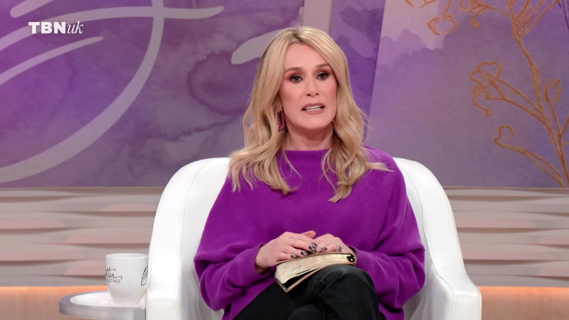 A woman with blonde hair, wearing a vibrant purple jumper, sits in a white chair. She holds an open book on her lap, her hands resting on its pages. The TBN UK logo appears in the top left corner.