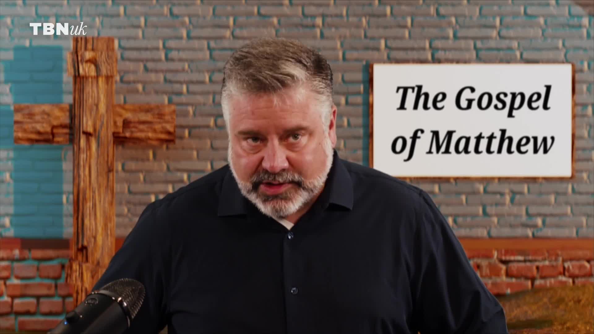 A man with a grey beard speaks directly to the camera, a wooden cross visible behind him. A sign on the wall behind him reads "The Gospel of Matthew."