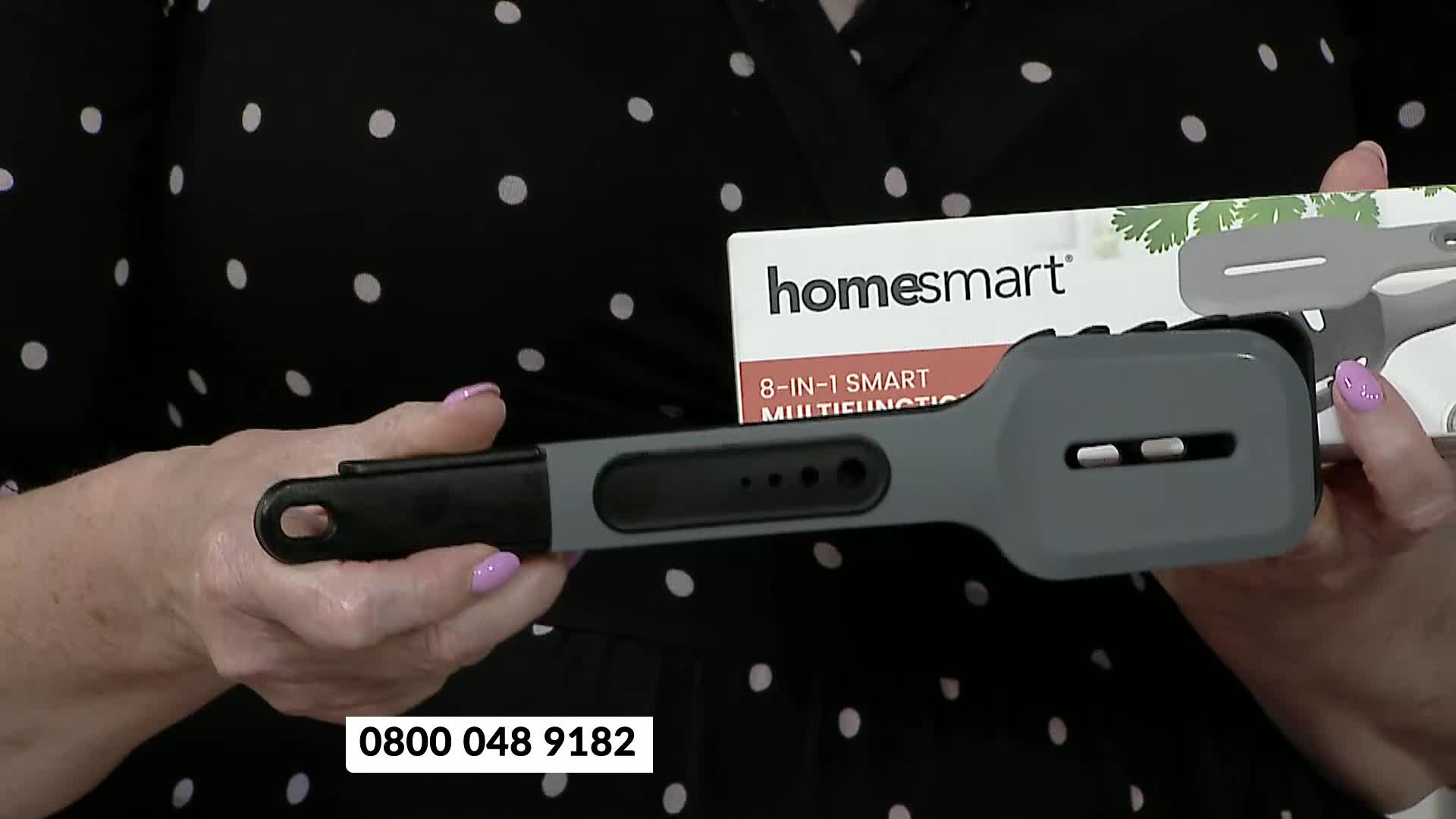 A woman in a polka-dot top is holding a gray kitchen tool, and a box of the same product is visible behind it. The phone number 0800 048 9182 is at the bottom of the screen.
