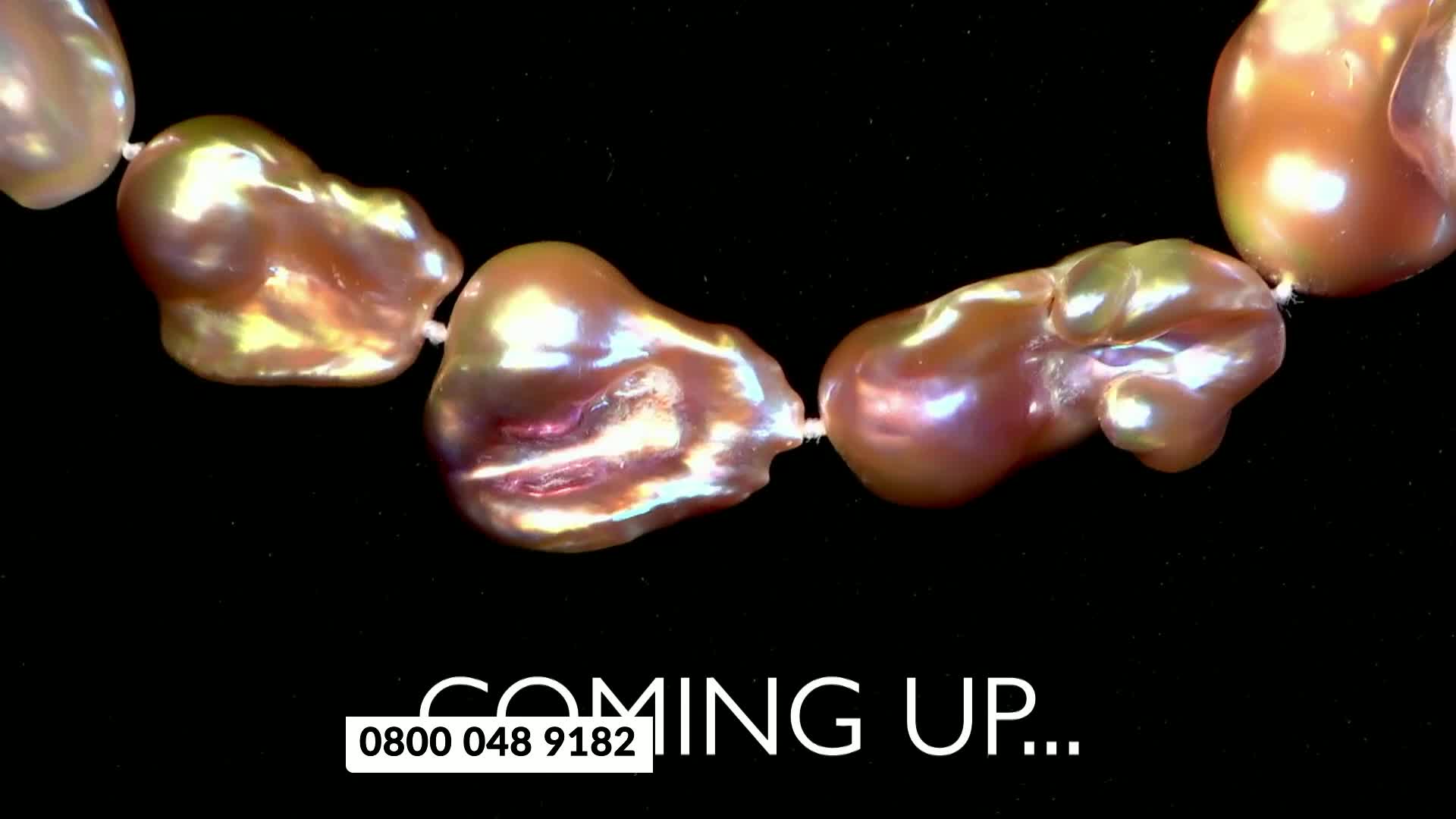 A string of large, irregularly shaped pearls gleams against a black background. The pearls shift in color, reflecting shades of pink, gold, and green.
