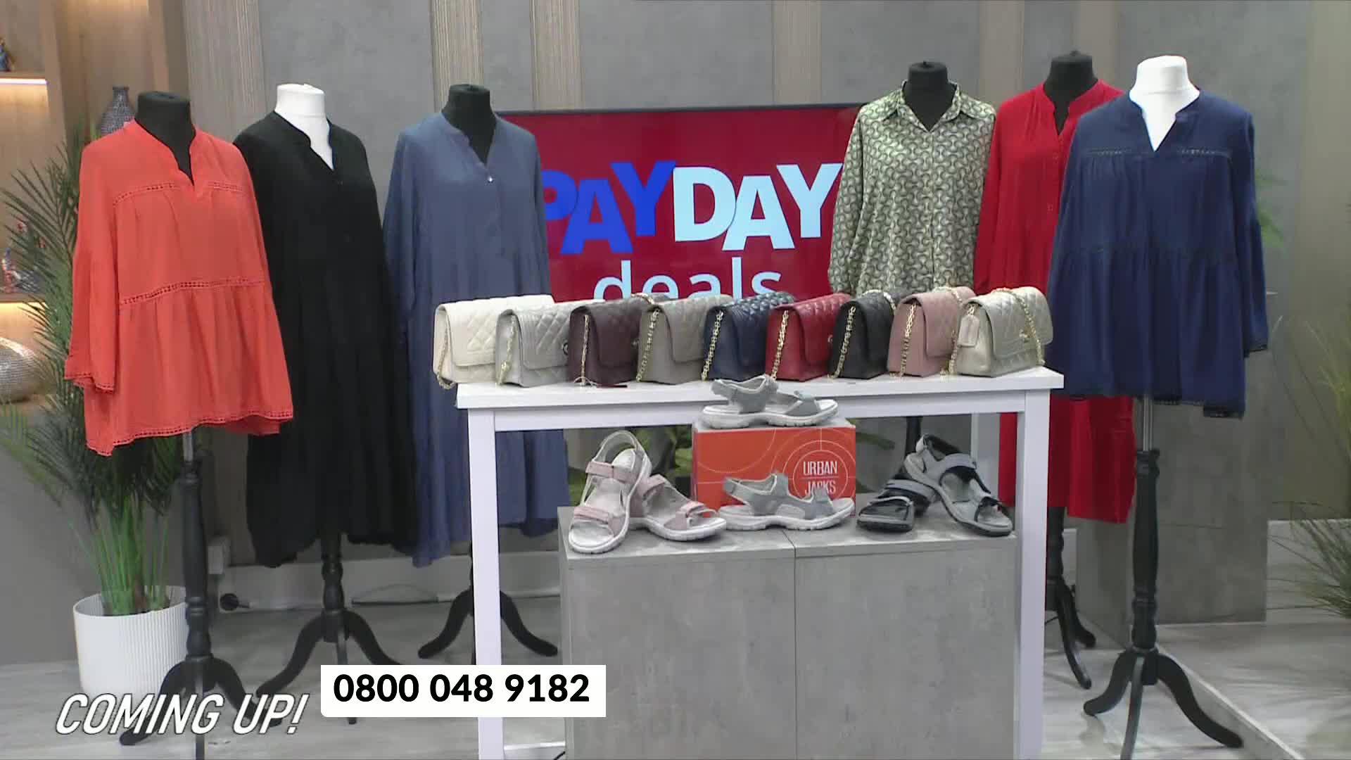 A display of clothing and accessories sits on a set, with a "Pay Day Deals" sign in the background. The items are arranged on mannequins and a small table, with sandals and handbags showcased.
