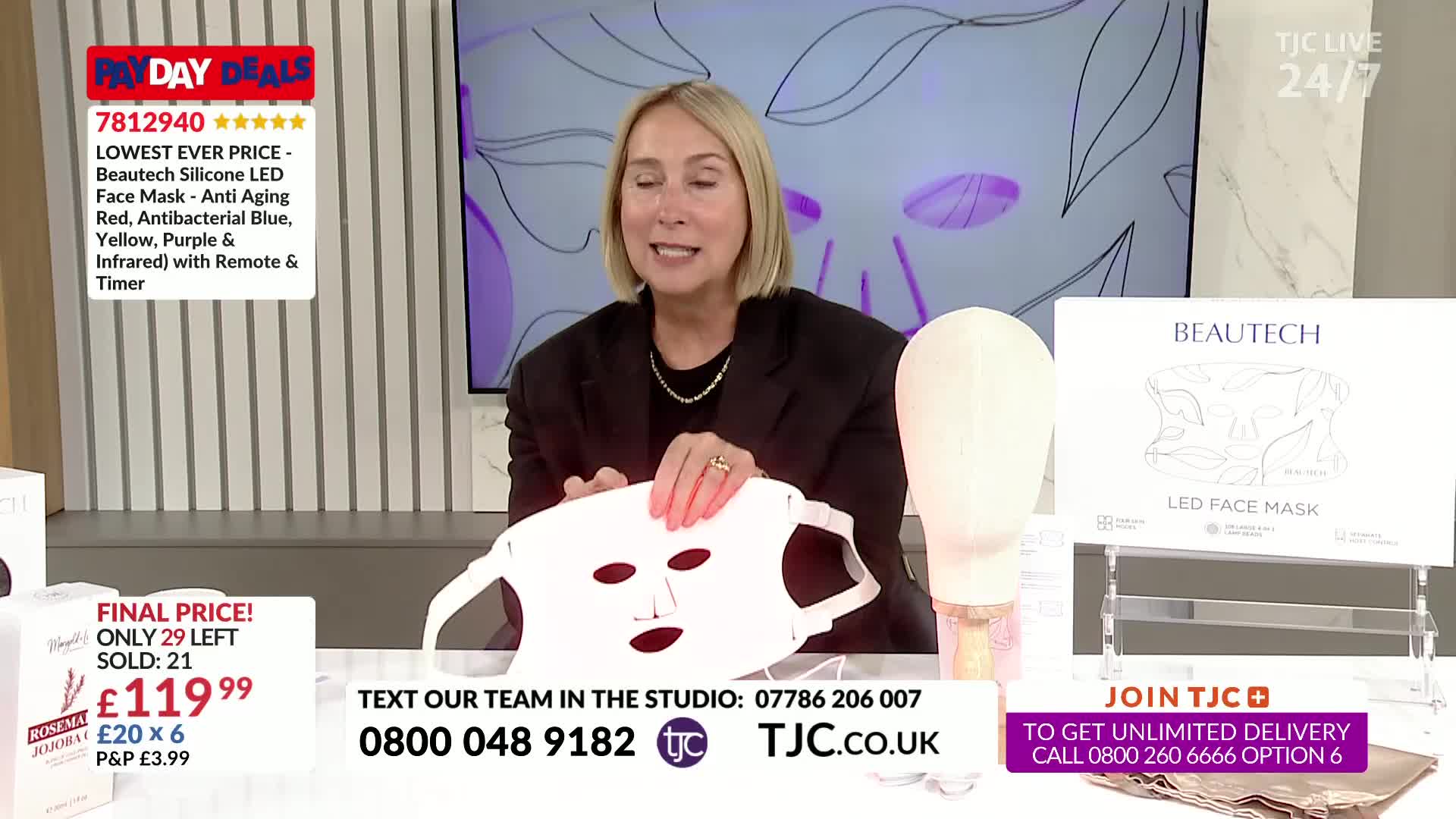 A woman on TJC is holding up a white, mask-like device, showing it to the camera. Behind her, a display advertises the product as an LED face mask.
