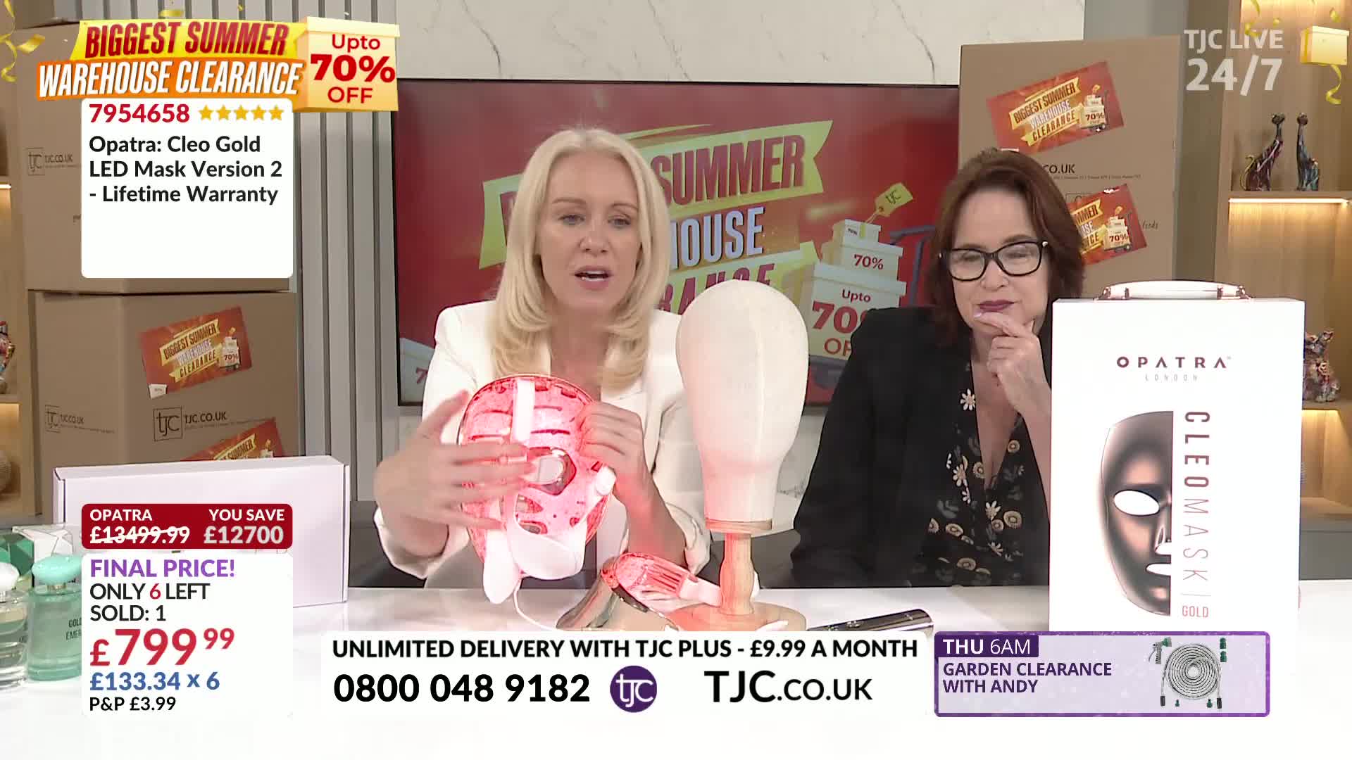 On a TJC set, a woman in a white jacket holds up a pink, futuristic-looking face mask, demonstrating it to the camera. Beside her, another woman in a black jacket looks on, with a box of the product resting on the table.

