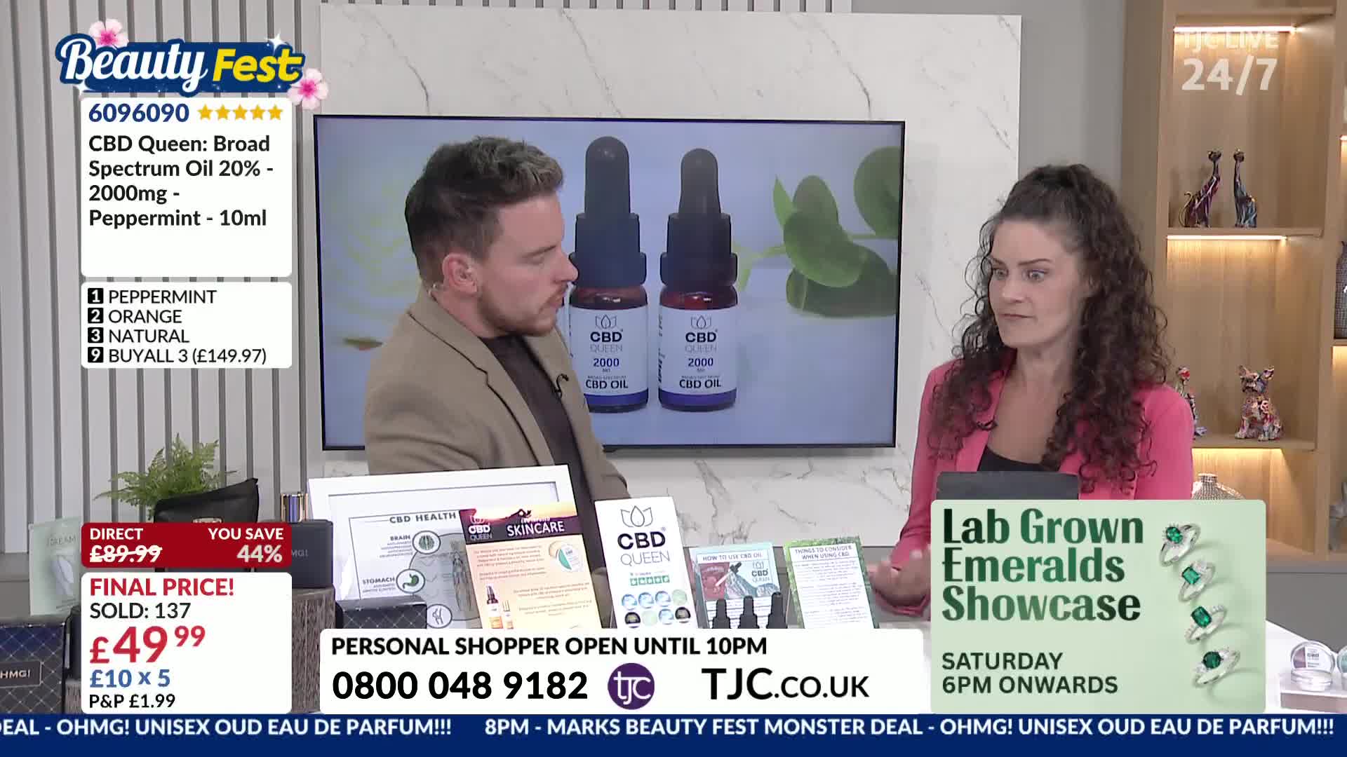 Two people are seated at a table, discussing CBD oil products on a TJC shopping channel. The woman gestures with her hands while the man looks towards the screen displaying the product.
