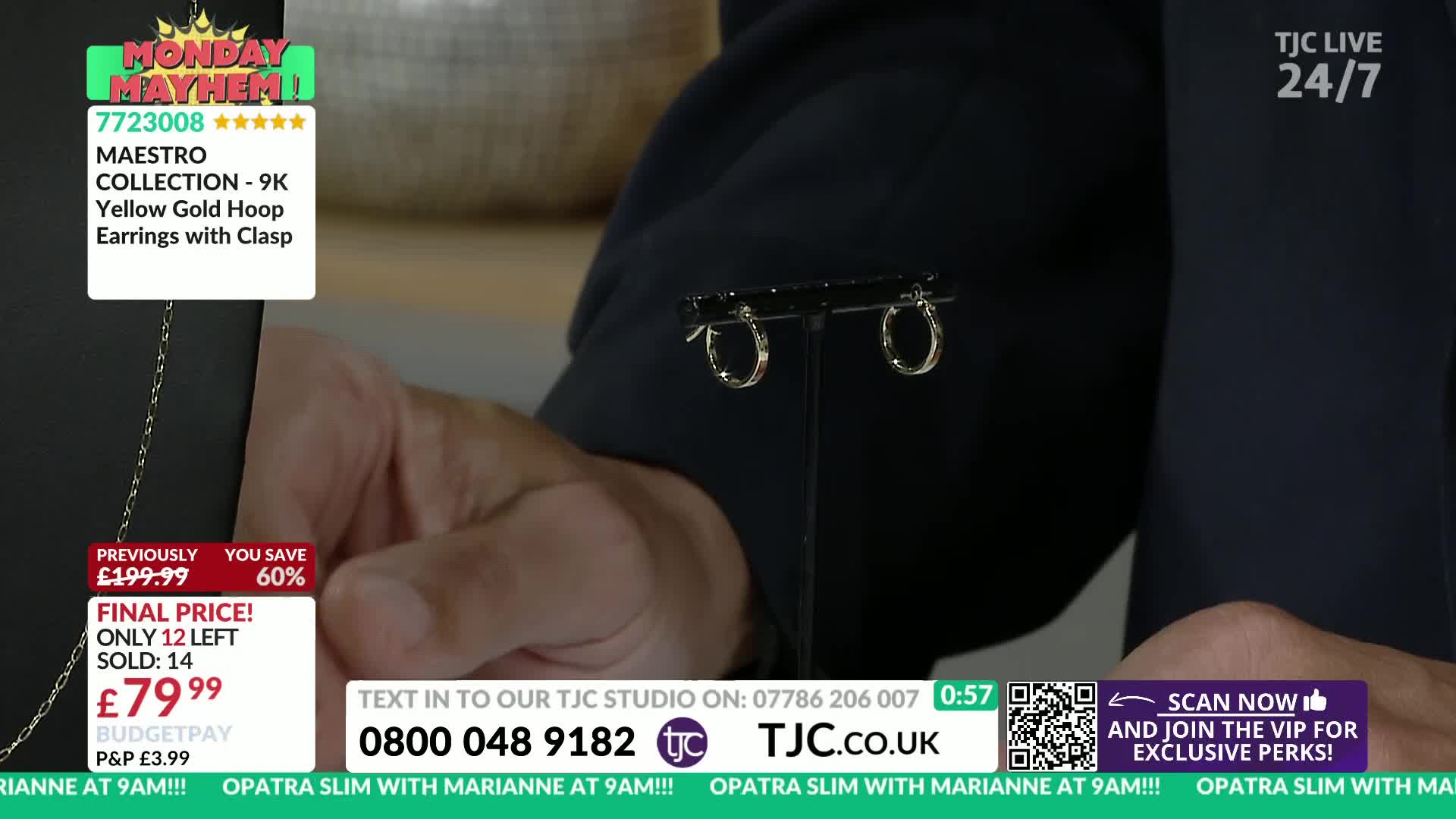 A pair of small gold hoop earrings sits on a black stand, held up by a person's hand. The TJC logo and a price tag are visible in the background.
