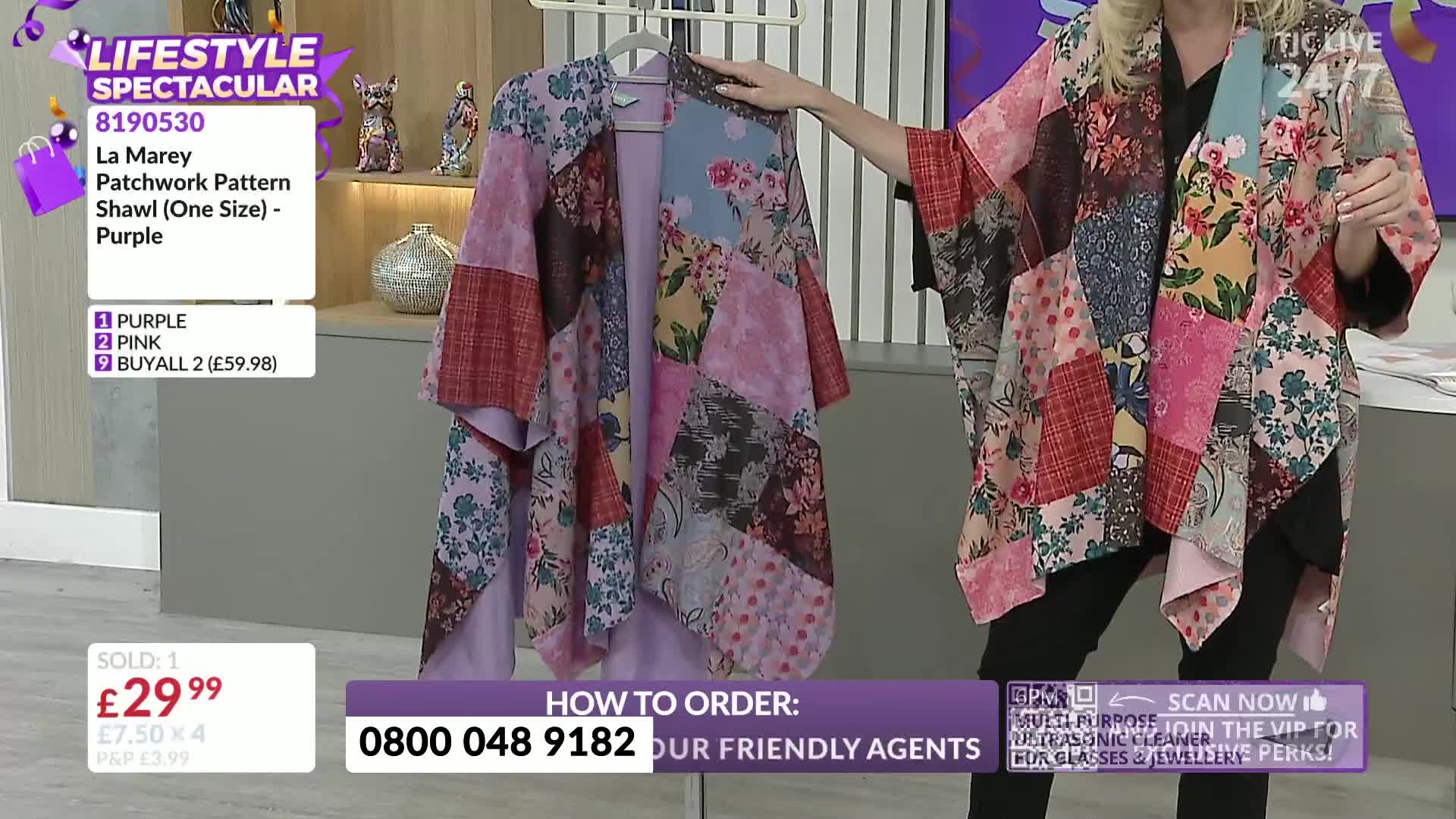 A woman on a TJC show in the United Kingdom holds up a patchwork shawl, showing it off as the camera zooms in. The garment, a mix of floral and patterned fabrics, hangs from a hanger on the left.
