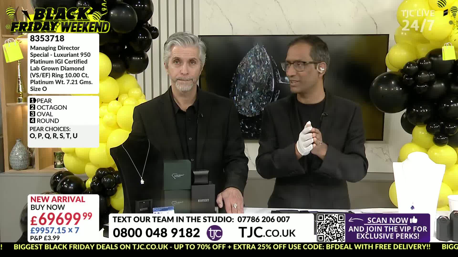Two men in black suits stand behind a table, with jewelry displayed in front of them. One man gestures with a gloved hand while the other looks towards the camera.
Two men in black suits stand behind a table, with jewelry displayed in front of them. One man gestures with a gloved hand while the other looks towards the camera.