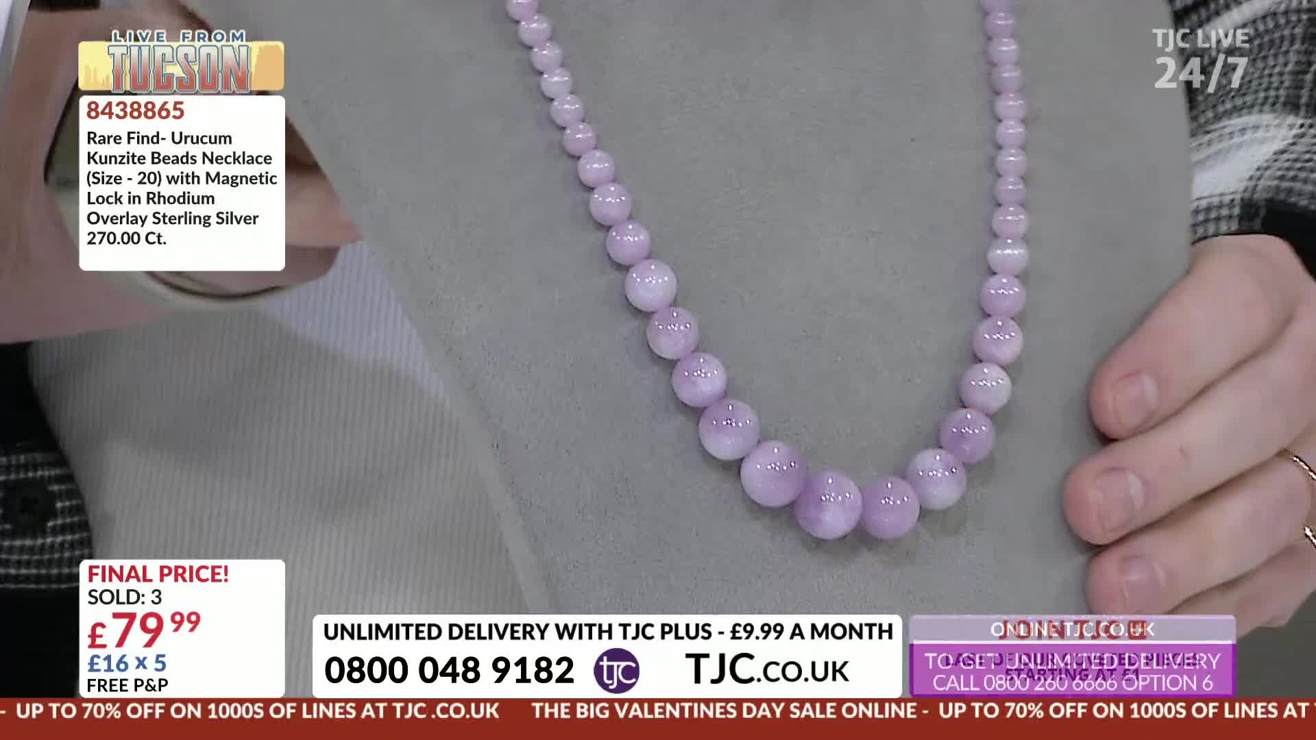 A strand of lilac beads, graduating in size, rests on a grey display. A hand with a ring gently adjusts the necklace, showing off its polished surface.