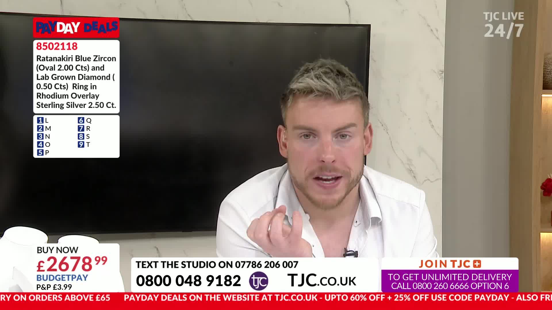 A man with styled blond hair is speaking animatedly, gesturing with his hands. A screen behind him displays product details for a Ratanakiri Blue Zircon ring.