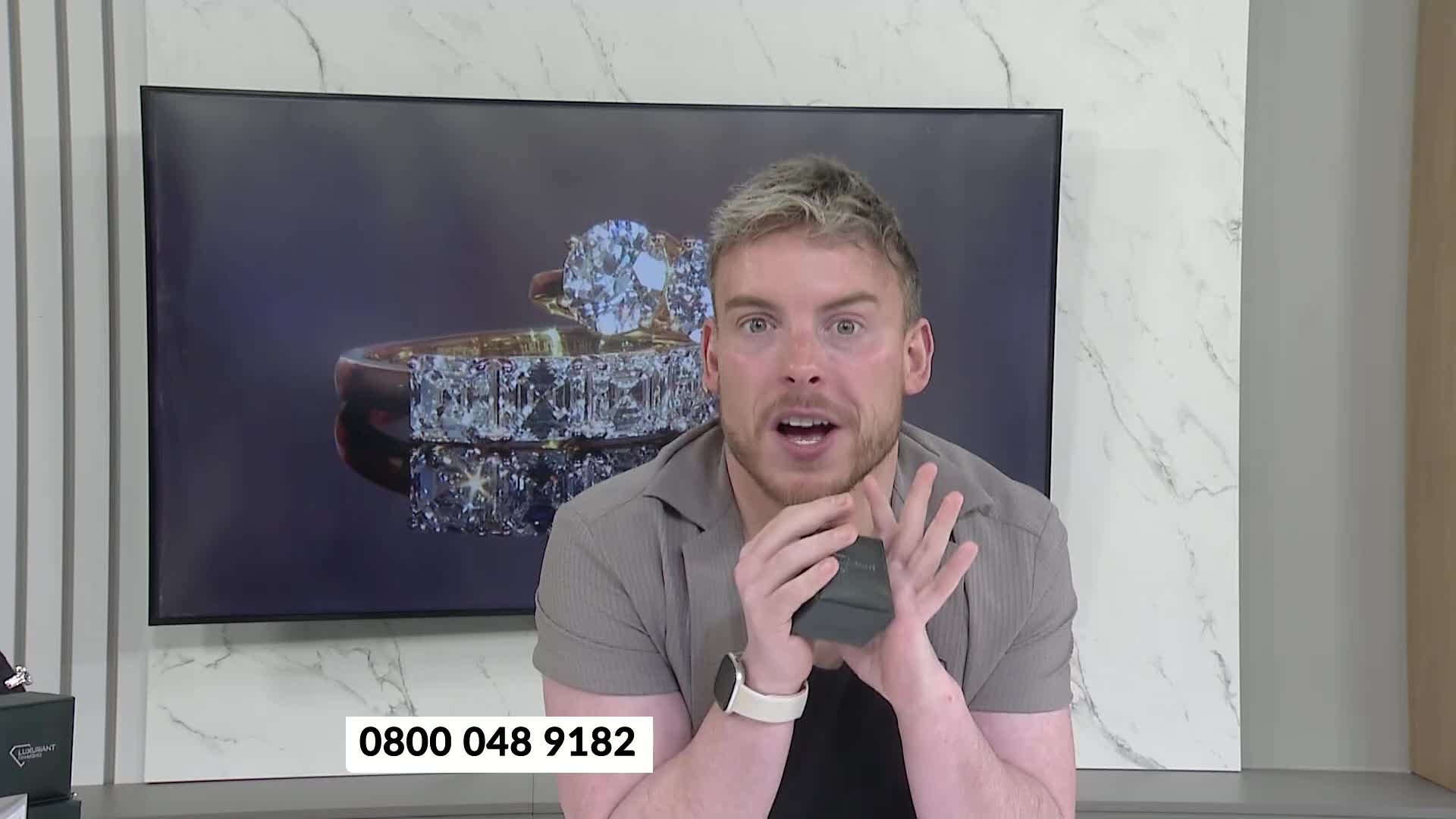 A man on TJC holds up a small, dark box, his mouth open in surprise. Behind him, a large screen displays two glittering diamond rings.
