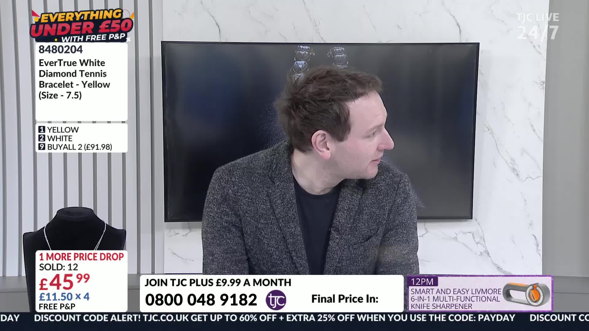 A man in a grey jacket is speaking, looking off to the side. Below him, a display shows a necklace on a black stand, with pricing information.