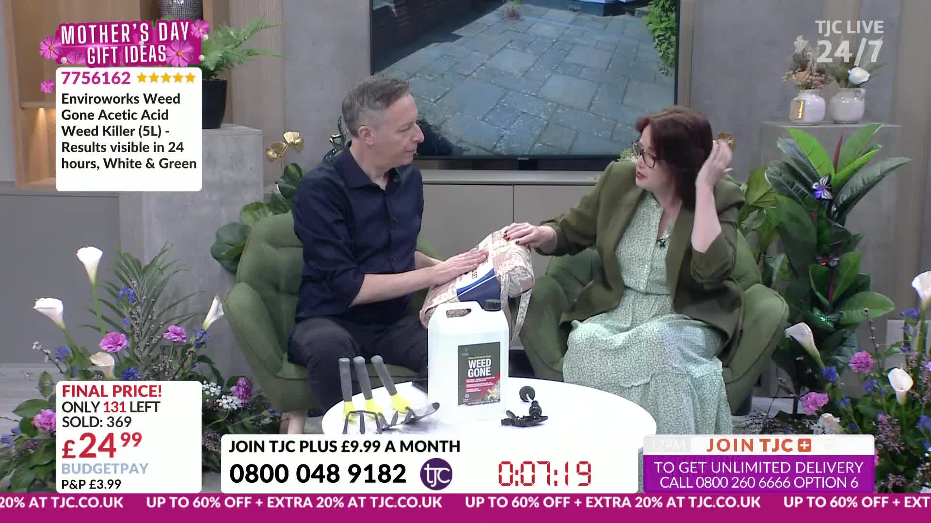 A man and woman are sitting on a sofa, discussing a large bottle of weed killer. The woman gestures towards the bottle as the man holds a patterned fabric.