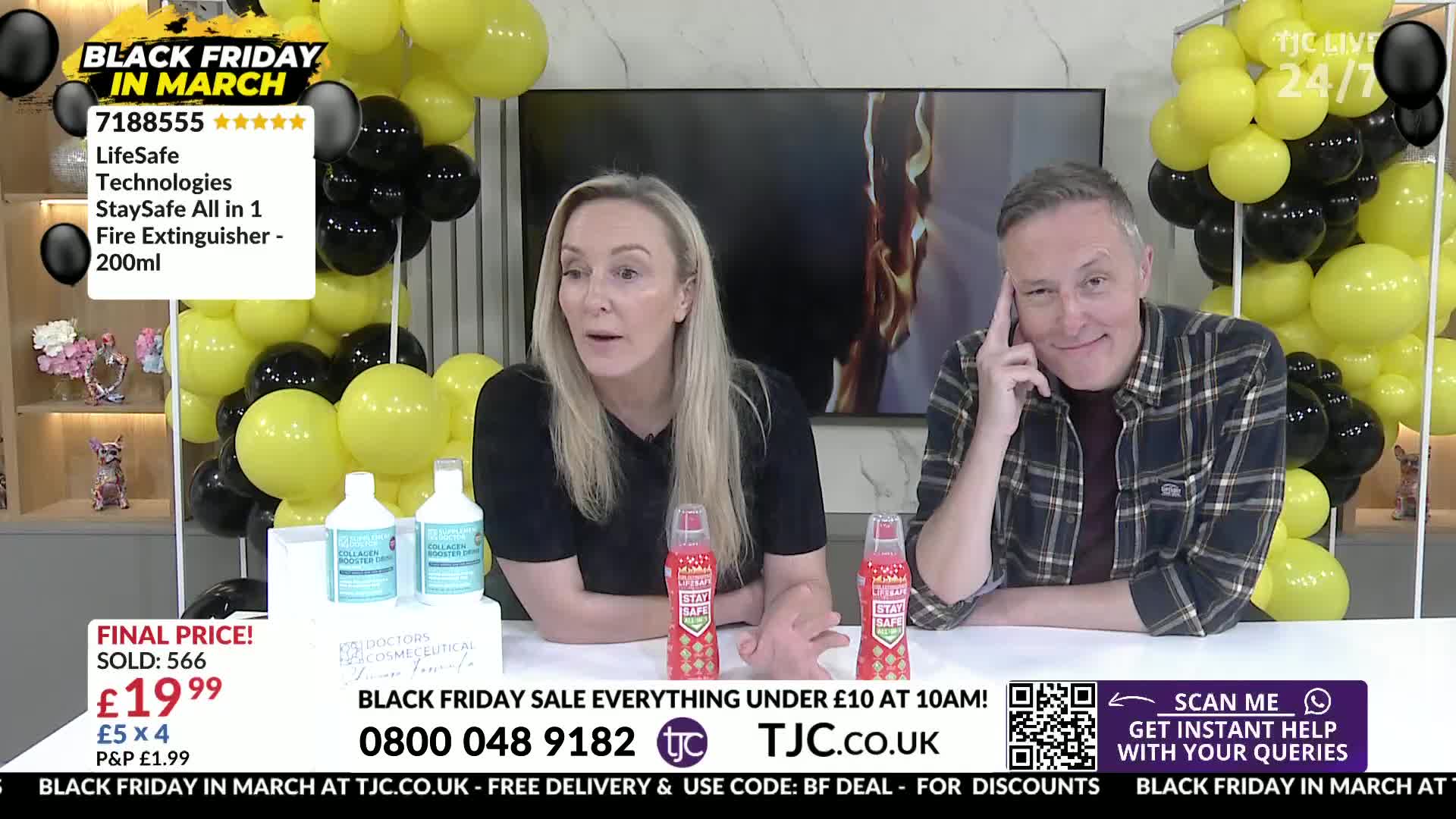 A woman with blonde hair gestures animatedly while a man in a plaid shirt smiles, both seated at a table. Behind them, a wall of yellow and black balloons frames a television screen displaying flames.