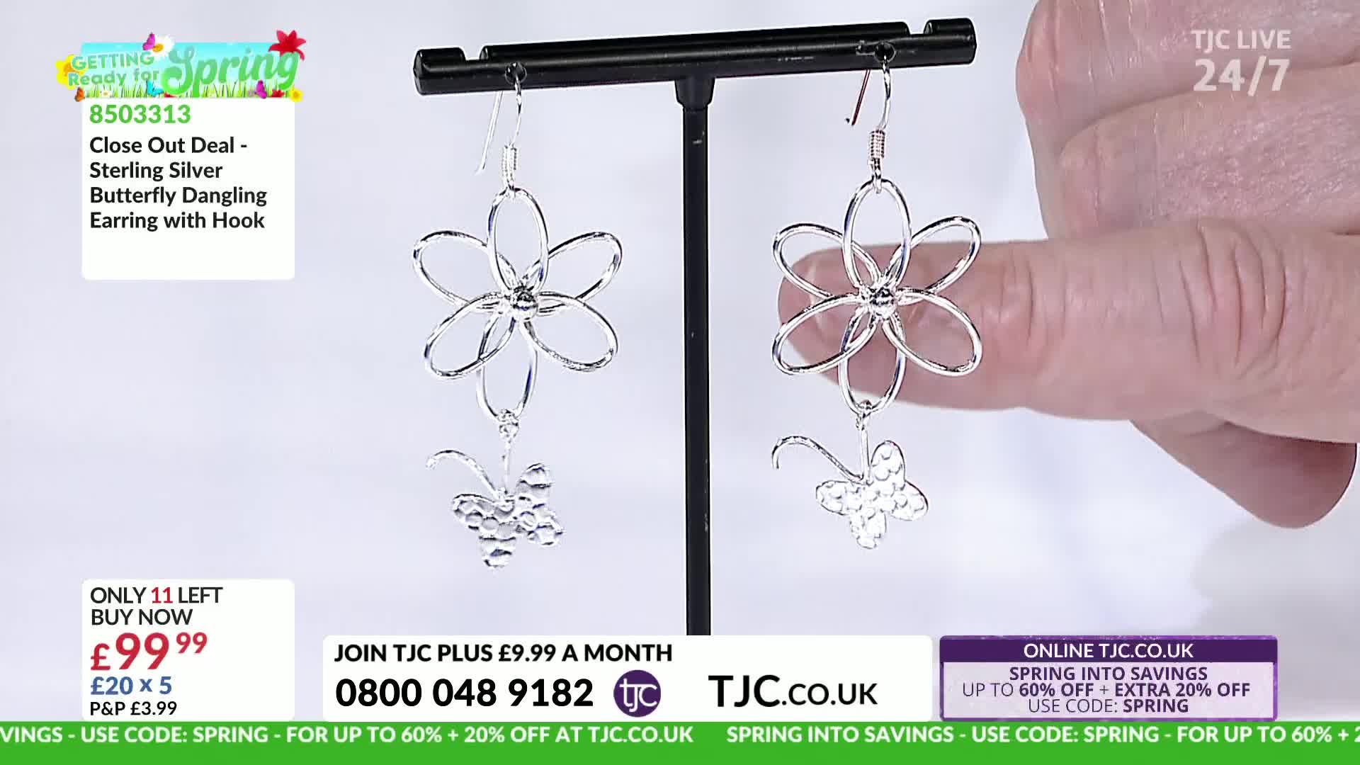 A hand gently holds a delicate silver earring, showcasing its flower and butterfly design. The earring dangles from a display stand, catching the light.