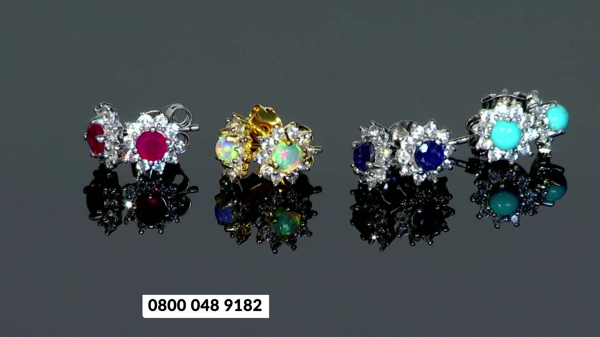 A row of four pairs of earrings sits on a reflective surface. The earrings feature various gemstones, including ruby, opal, sapphire, and turquoise, surrounded by small clear stones.