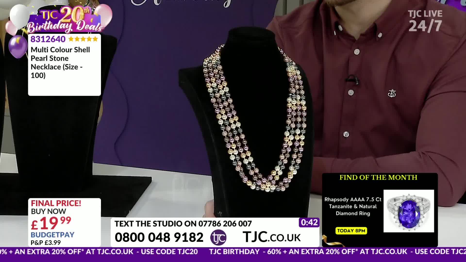 A multi-strand necklace of colourful shell pearls rests on a black stand. A man in a maroon shirt stands behind it, with a TJC logo visible.