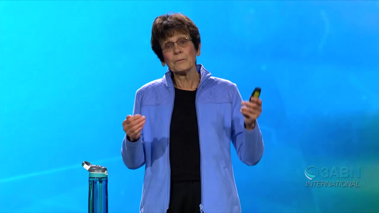 A woman in a blue jacket gestures with her hands as she speaks, holding a small, yellow-and-black object. A water bottle sits nearby on the floor, and the 3ABN International logo is visible.
