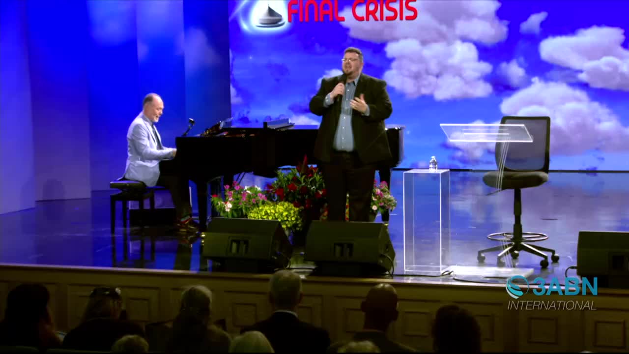 A man in a suit gestures with his hands as he speaks into a microphone on a stage. Beside him, another man plays the piano, their performance framed by a backdrop of a cloudy sky.
