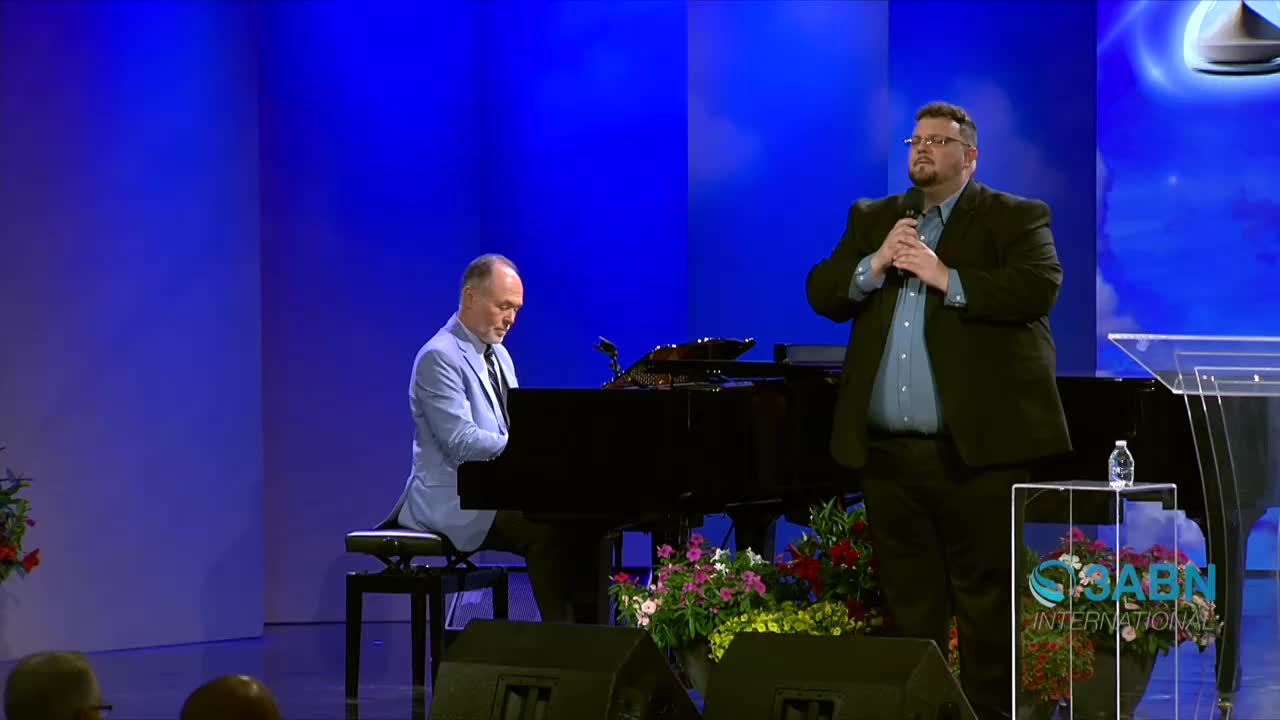 A man in a suit holds a microphone, speaking from a podium. Beside him, a pianist in a light blue jacket plays a grand piano.
