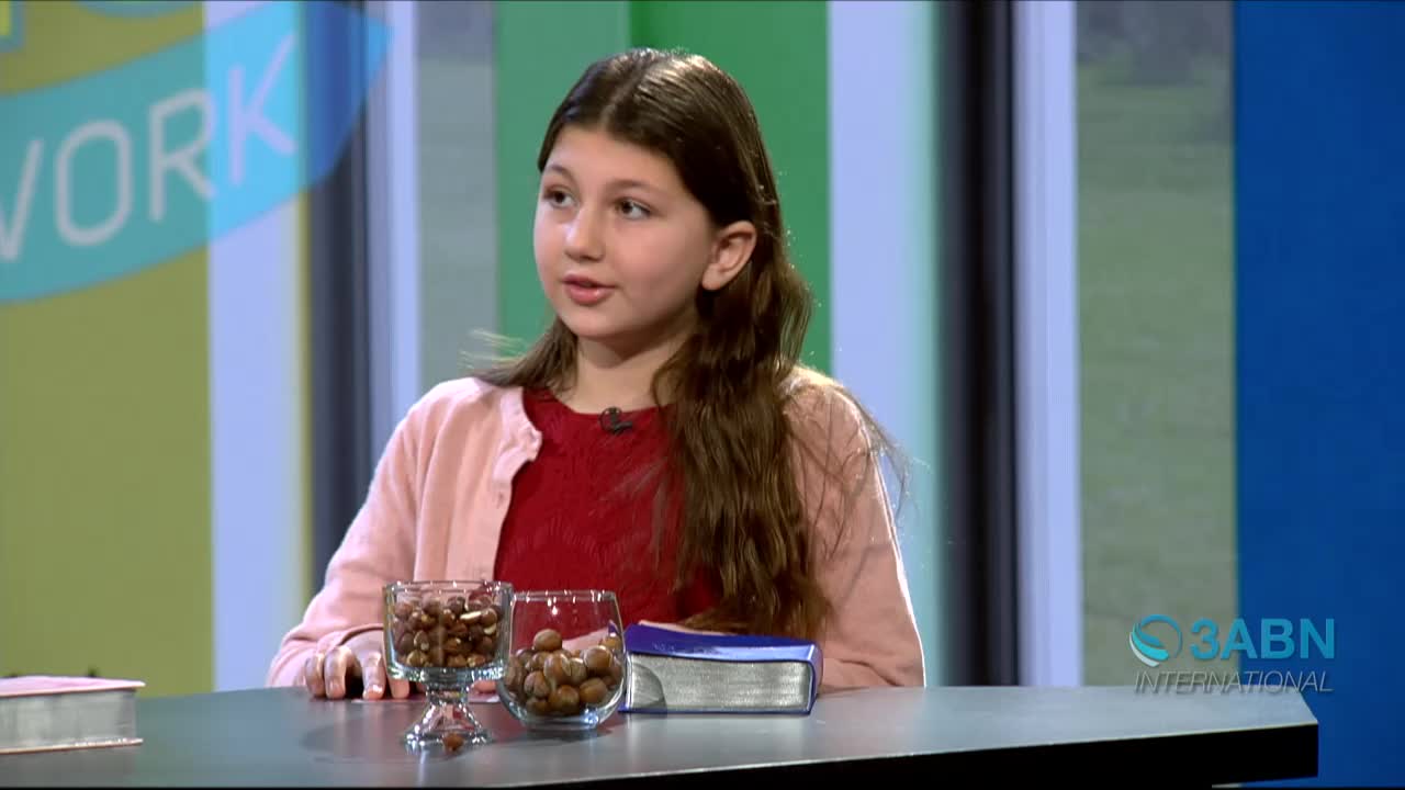 A young girl in a pink cardigan and red shirt sits at a table, speaking on a 3ABN International set. Two glass bowls filled with nuts and a closed book rest in front of her.
