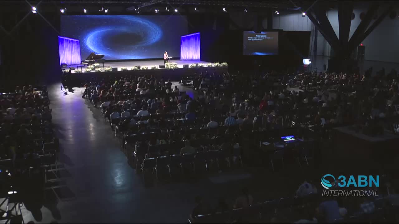 A speaker stands on a stage, addressing a large crowd in a vast hall. The audience is seated in rows, listening intently as the presentation unfolds.
