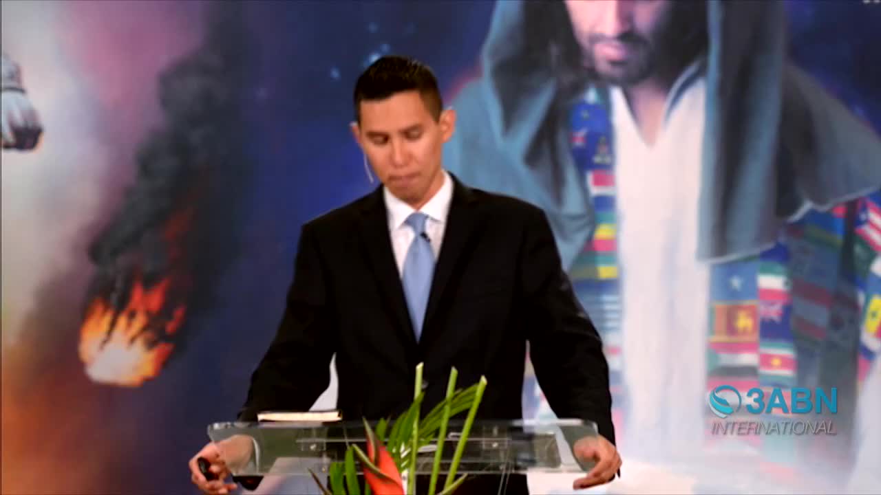 A man in a suit stands at a clear podium, speaking in front of a backdrop featuring a depiction of Jesus and a fiery scene. The 3ABN International logo is visible in the lower right corner.
