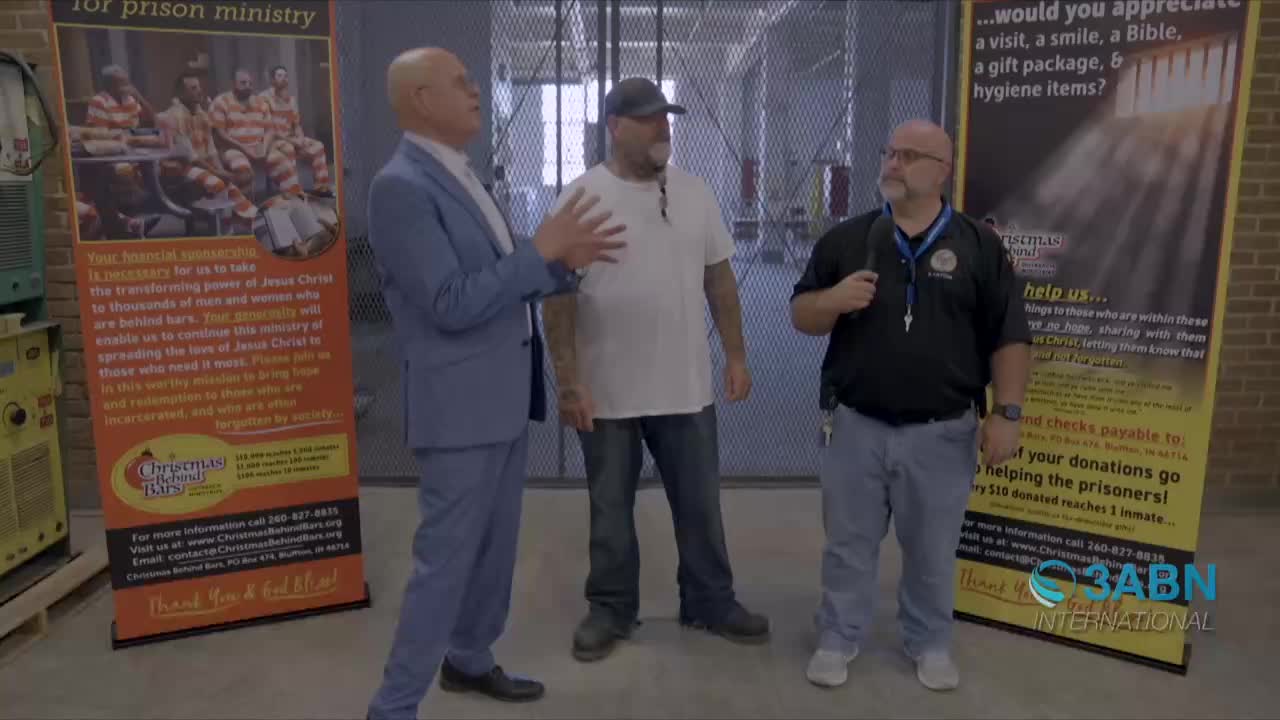 A man in a blue suit gestures with his hands as he speaks to a man in a white t-shirt and a corrections officer. Behind them, a metal gate and banners for a prison ministry are visible, with the 3ABN International logo in the corner.
