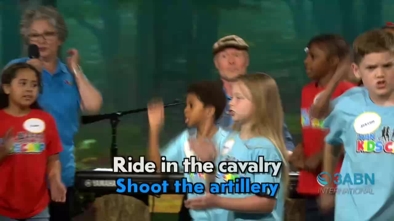 Children, wearing name tags, sing into microphones on a stage, while an older woman with glasses holds one as well. The words "Ride in the cavalry, Shoot the artillery" are displayed on the screen.
